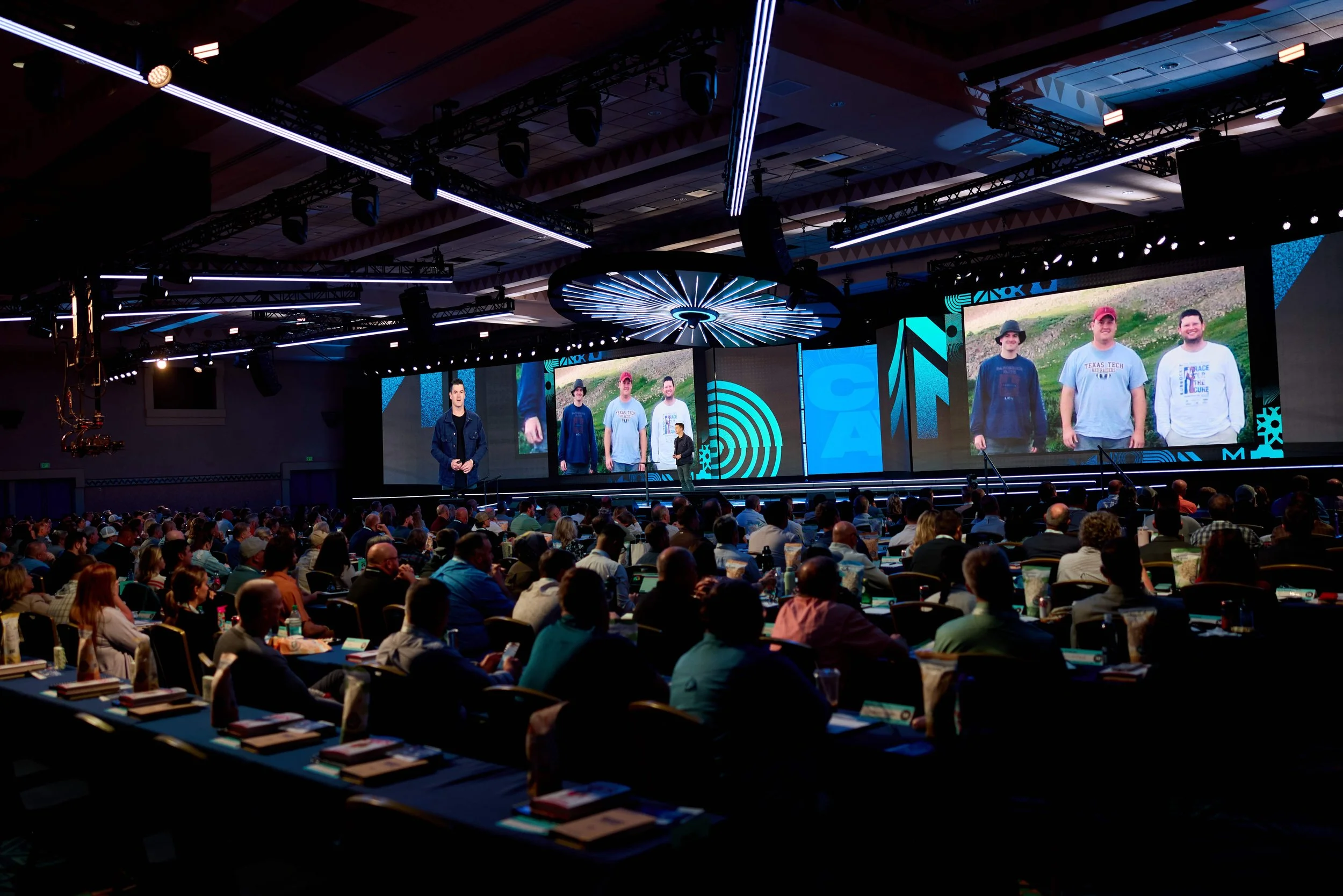 A large conference held in a spacious auditorium with many attendees seated at tables. A stage at the front displays large screens showing photos of three young men outdoors, with a modern ceiling light fixture above the stage.