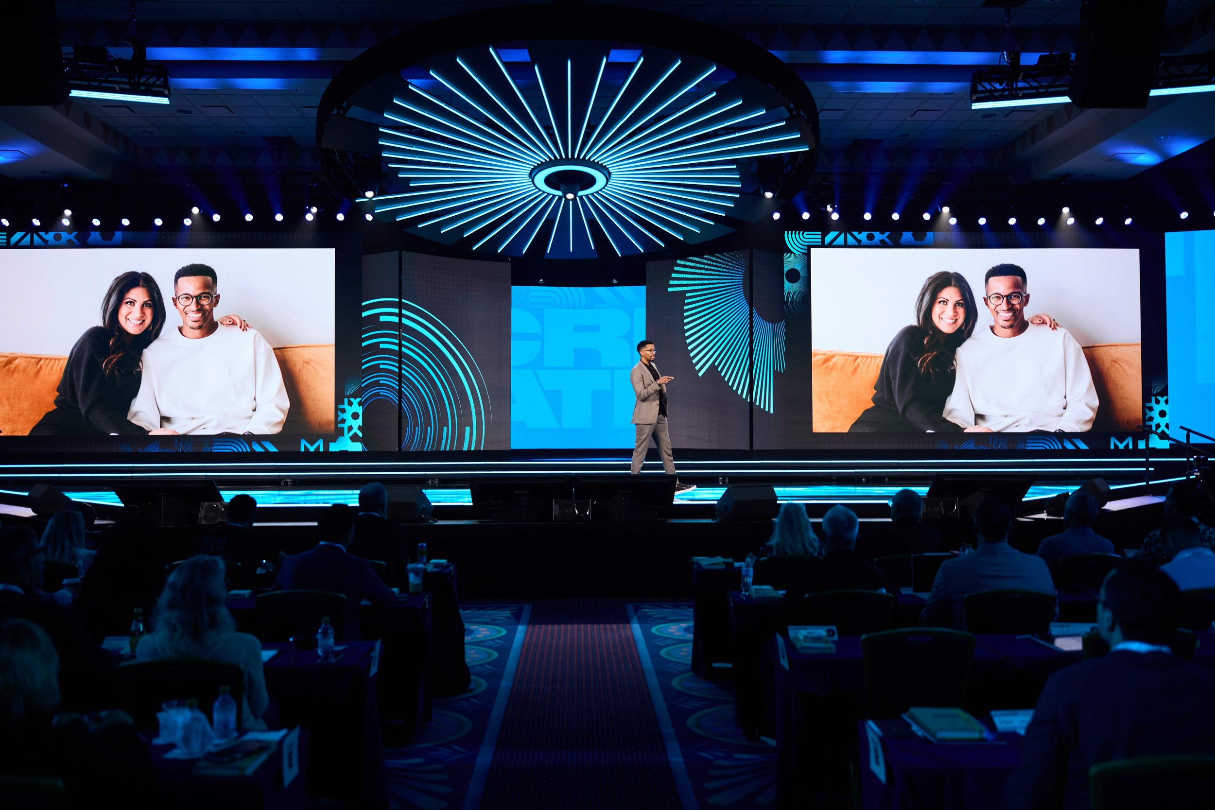 A man in a gray suit is giving a presentation on a stage with large screens displaying images of two people sitting together, smiling. The stage has blue lighting and a modern ceiling fixture, with an audience seated in front.