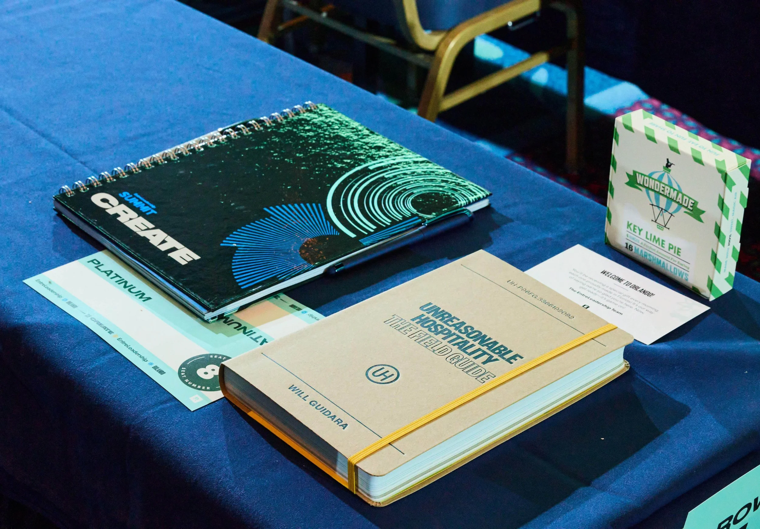 Gifts that Platinum tier attendees get at their seat. It includes the Summit Create workbook, a Platinum name tent, a welcome card, branded pen, Will Guidara's book, and a snack.