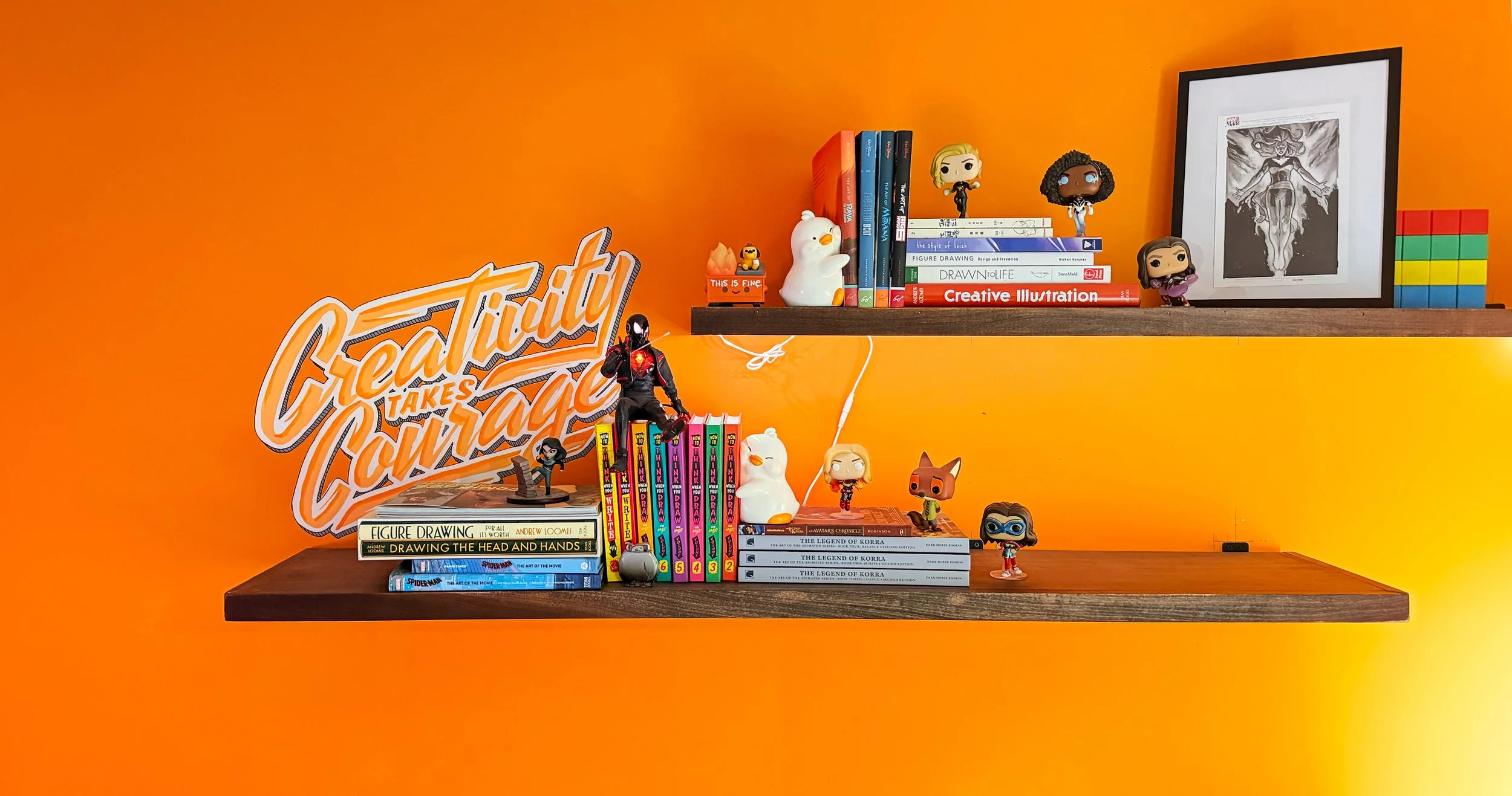 Orange wall with two dark brown floating shelves filled with books, figurines, and artwork. The lower shelf has a sticker with the phrase "Creativity takes Courage." The shelves display various books, including art, drawing, and fiction, along with Funko Pop figures, small toys, and framed black-and-white artwork. There is a sign on the left side of the lower shelf that says "Figure Drawing" and a Spider-Man comic book.