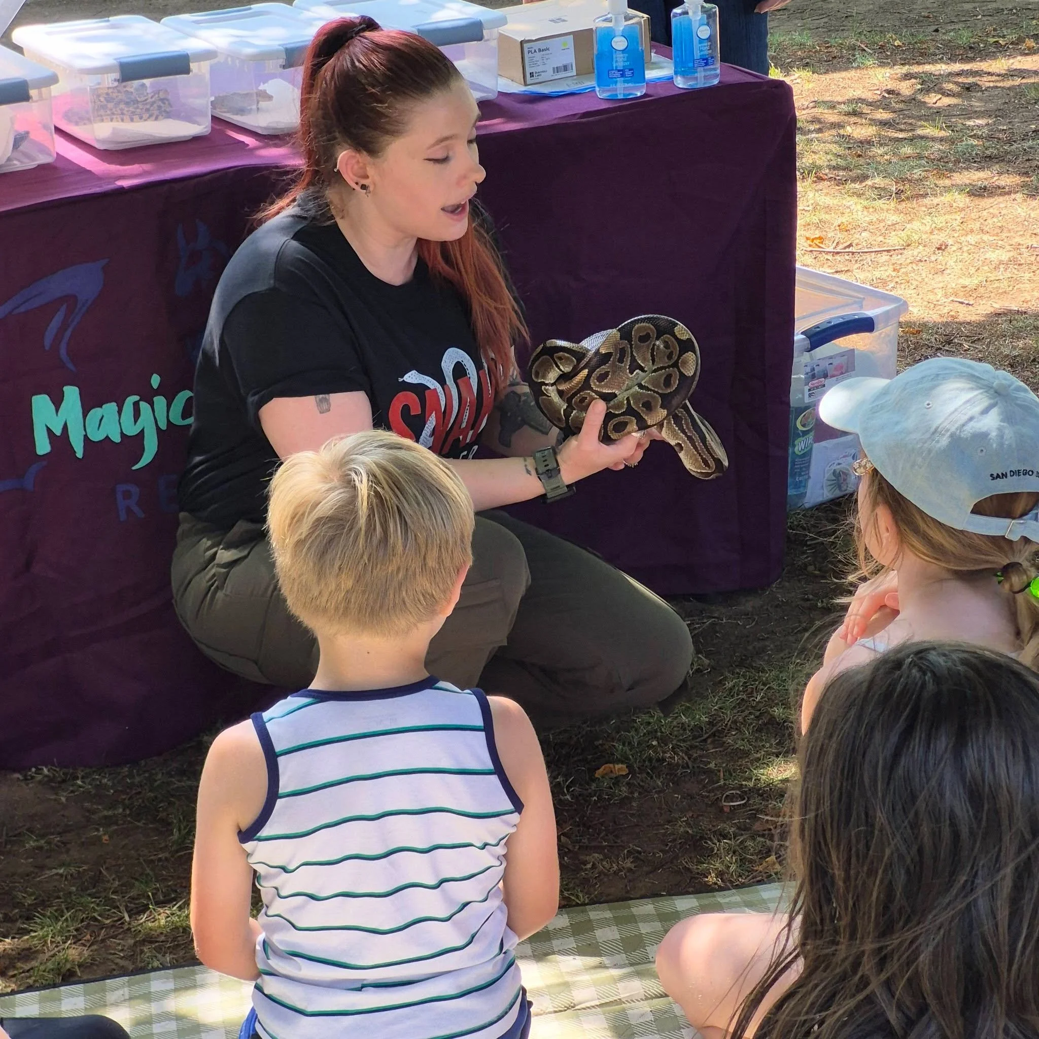 A woman with long red hair showing a snake to children at an outdoor event, with tables displaying various items and bottles of hand sanitizer in the background.
