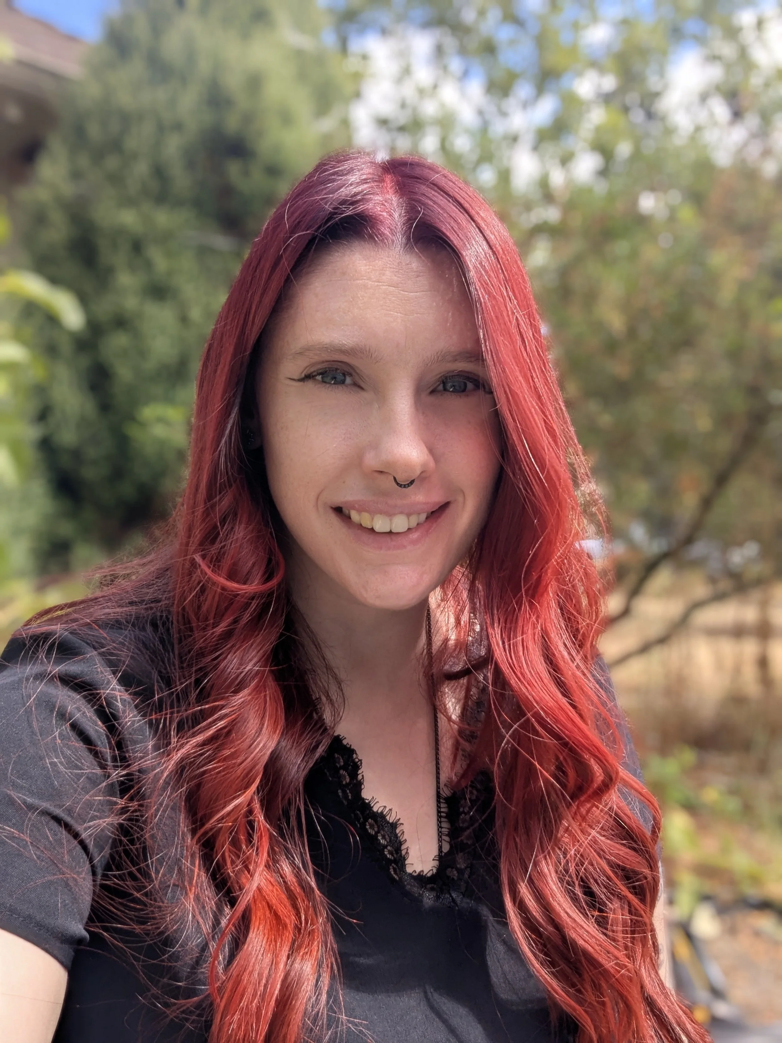 A smiling woman with red hair and a septum piercing taking a selfie outdoors, with trees and a house in the background.