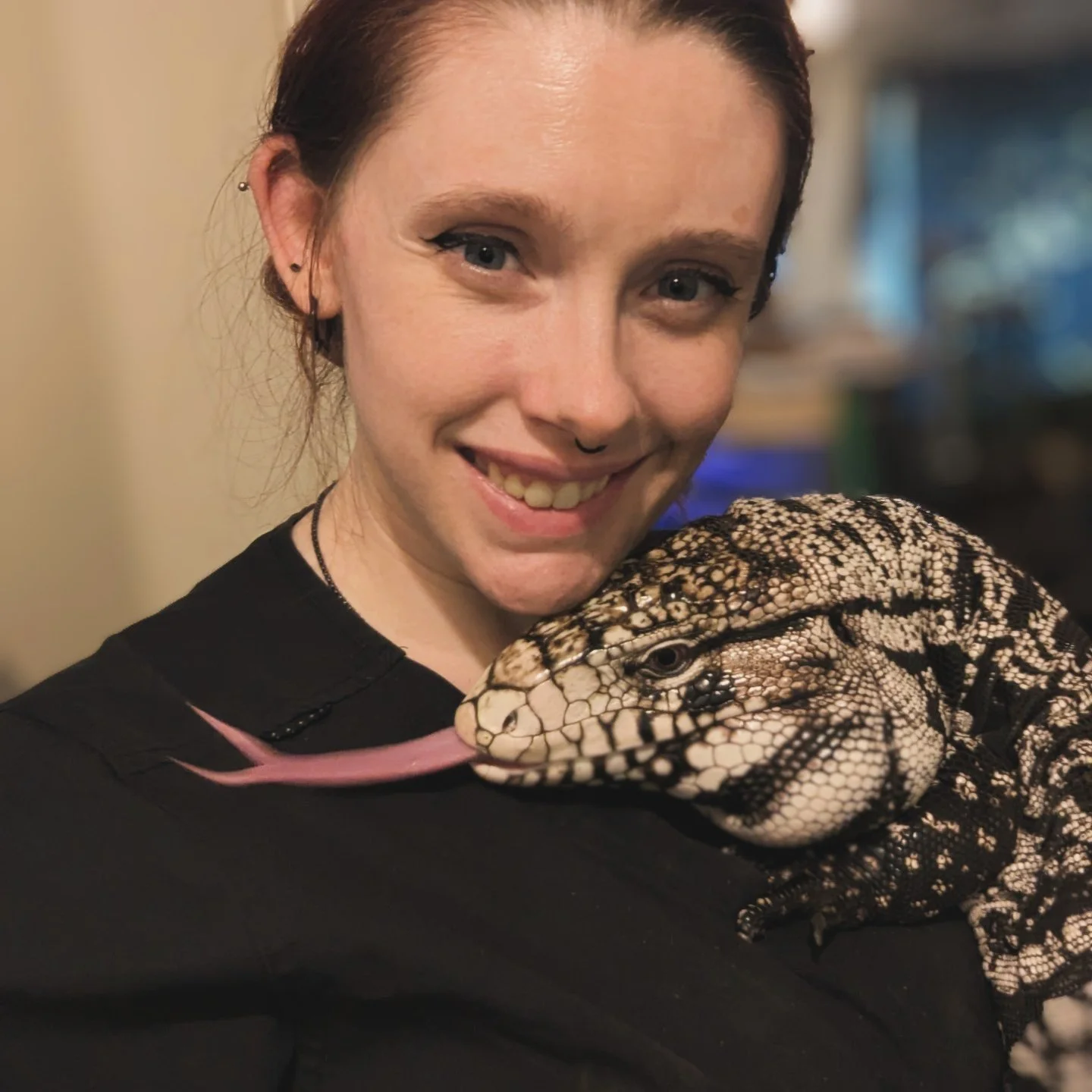 A woman with short red hair and piercings is smiling and holding a large black and white lizard with a long tongue near her face.