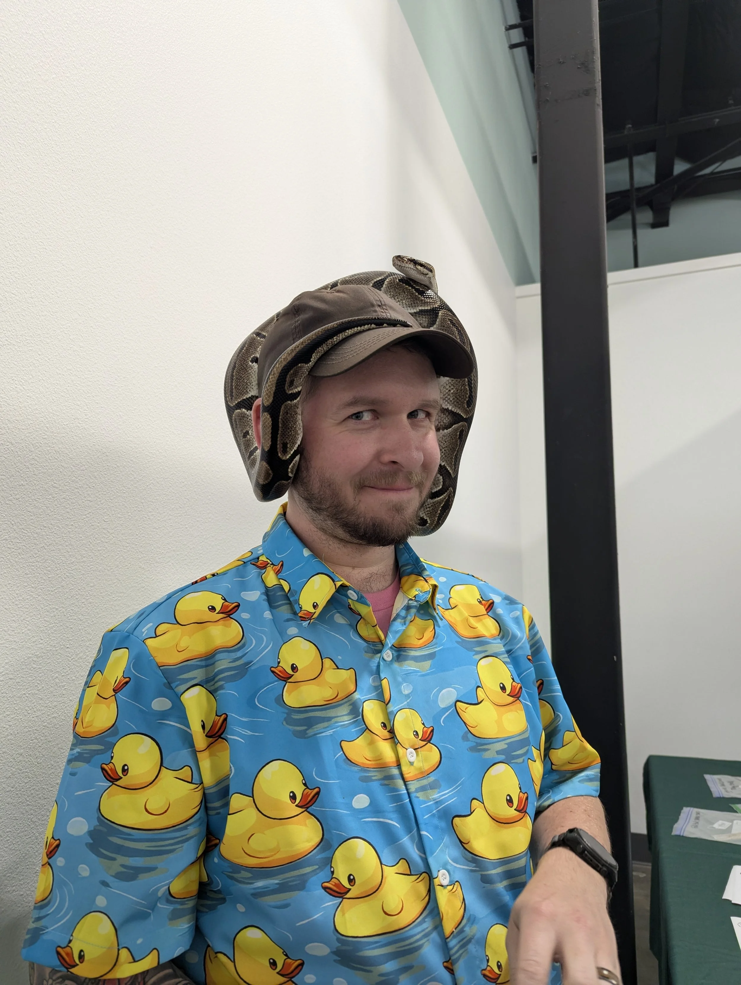 A man wearing a blue Hawaiian shirt decorated with yellow rubber ducks, a brown cap, and a snake draped around his shoulder. He is smiling and pointing with his right hand.