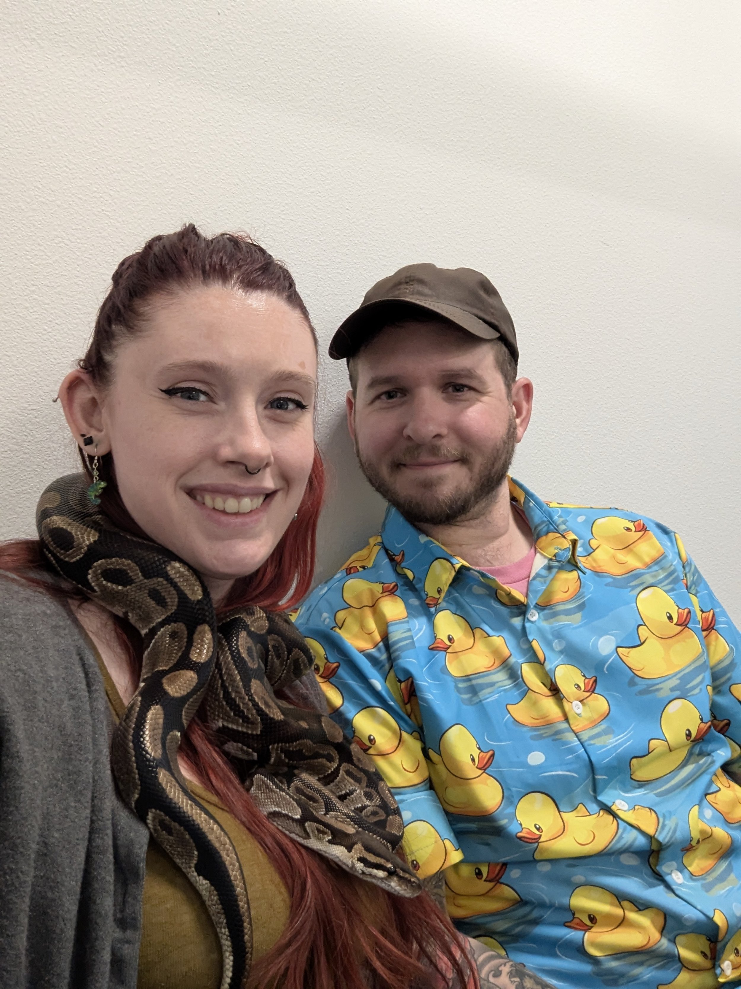 Two people smiling, a woman with red hair, black eyeliner, and earrings, and a man wearing a brown cap and a blue shirt with yellow rubber duck prints, standing against a plain white wall.