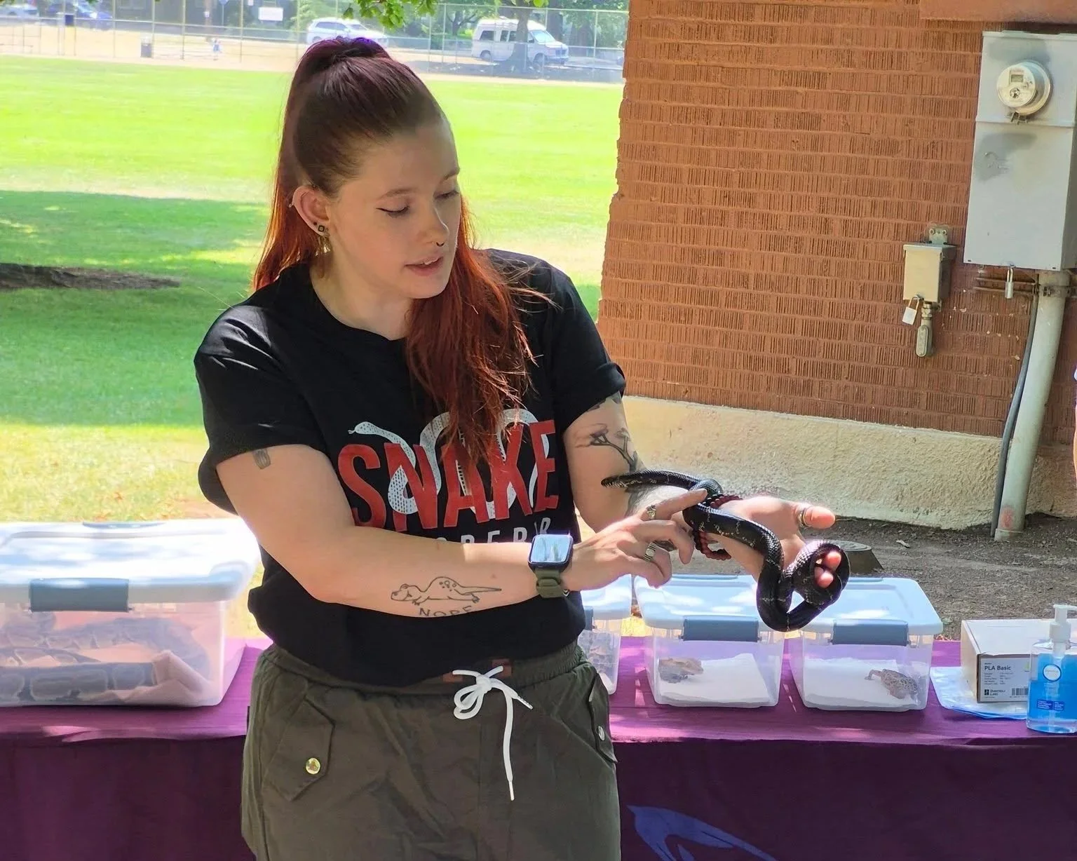 A woman with long red hair, tattoos, and a black t-shirt holding a black snake outdoors under a covered area with a brick wall and green field in the background.