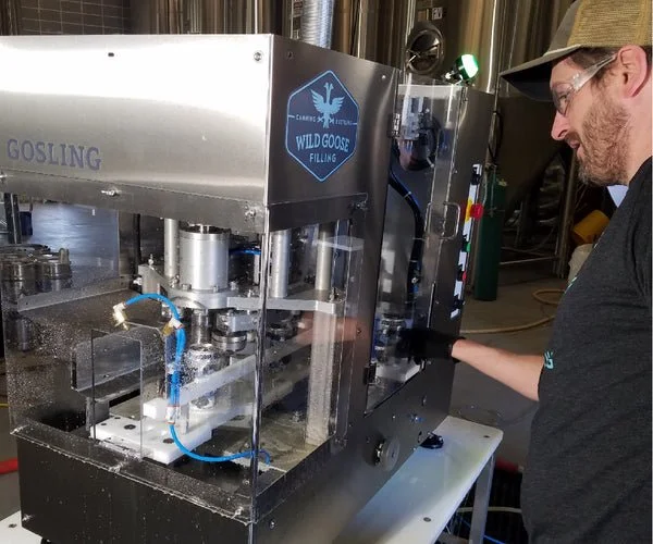 A man operating a Wild Goose filling machine in a brewery or distillery, filling bottles with liquid.