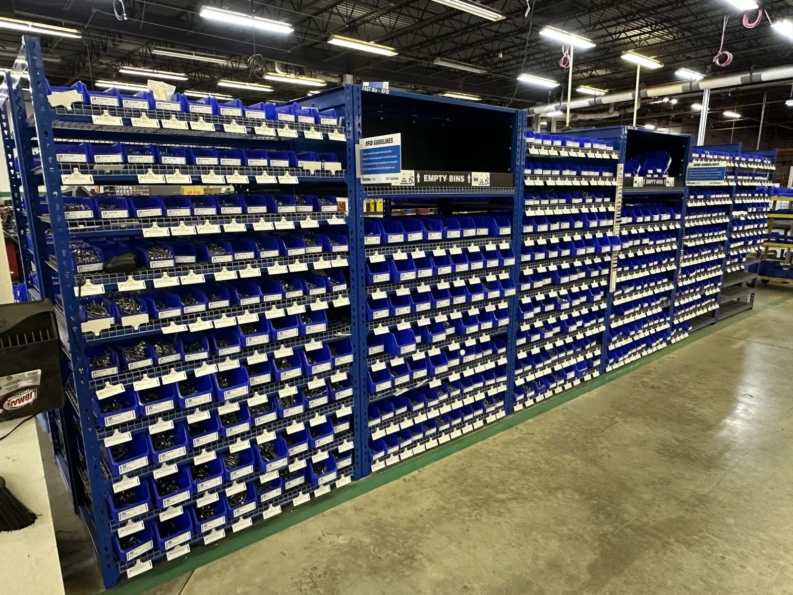 A large blue warehouse storage rack filled with small plastic bins containing various small hardware parts, with signage indicating return and empty bins.