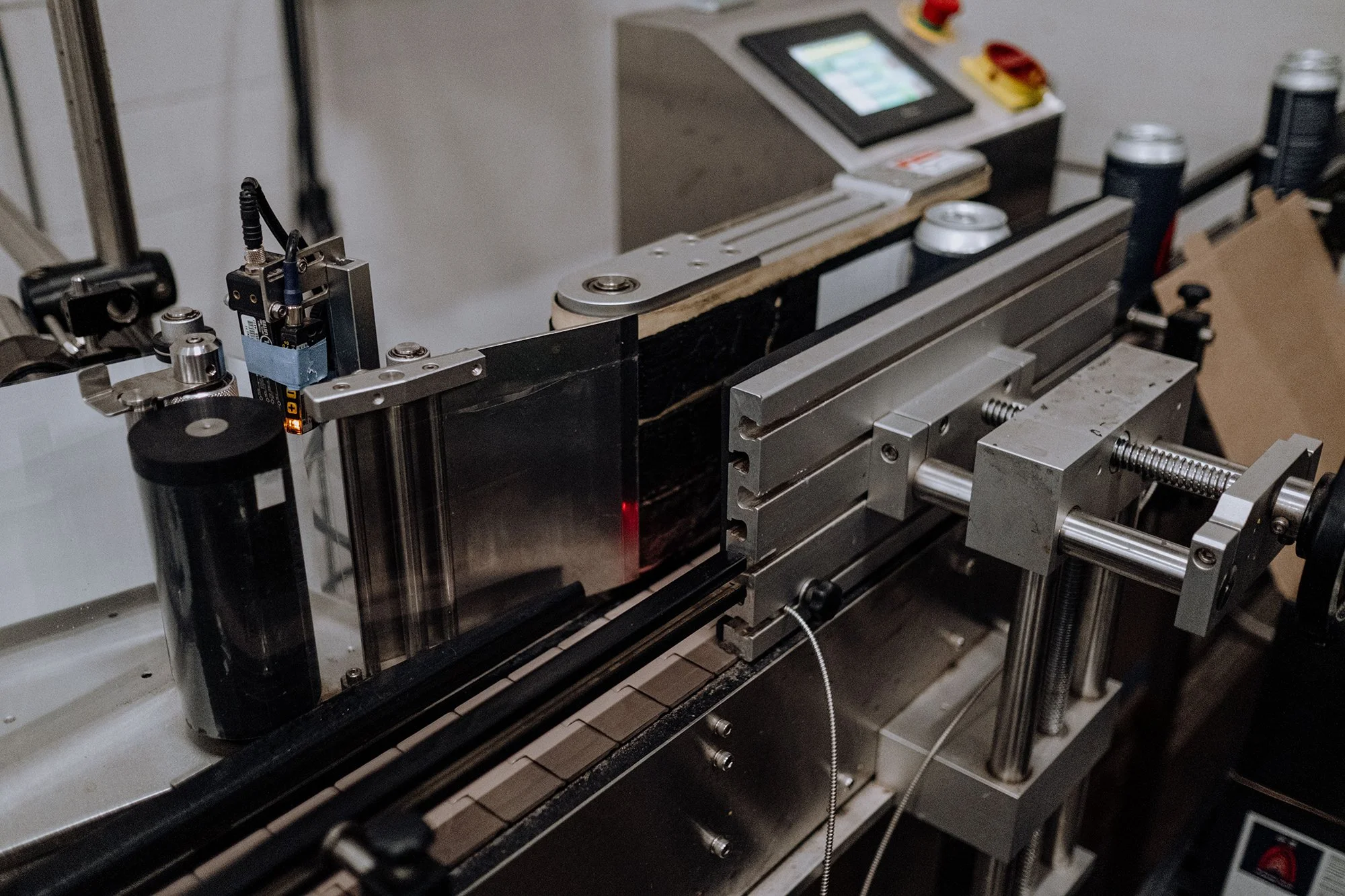 Close-up of a mechanical or industrial machine with metal components, rollers, and a digital control panel in the background. Two cans are placed on the machine.