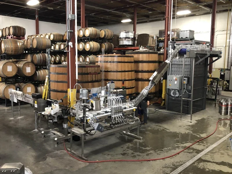 Industrial canning machinery in a warehouse with stacked barrels in the background