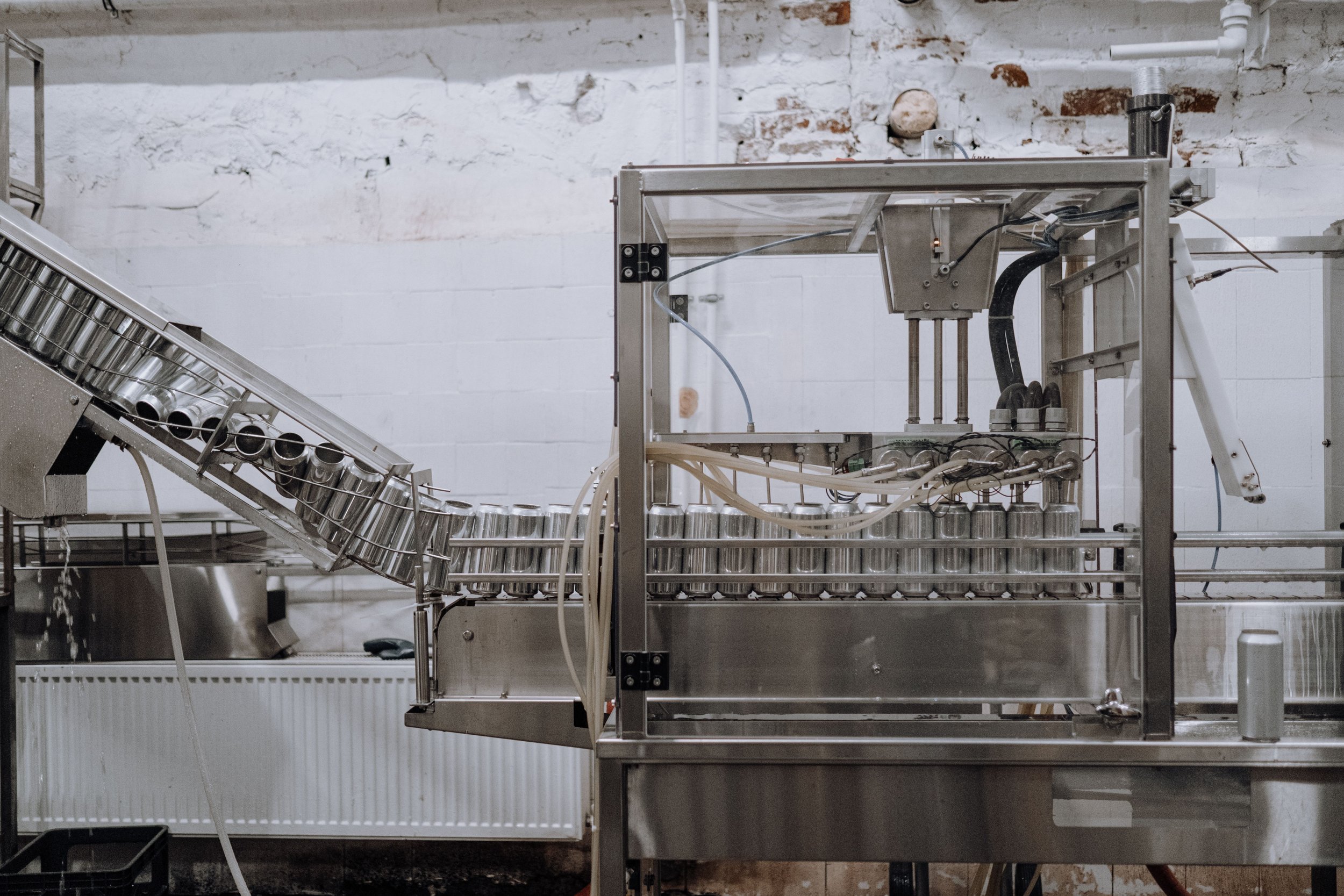 A stainless steel industrial machine with a conveyor belt system, set against a white brick wall background.