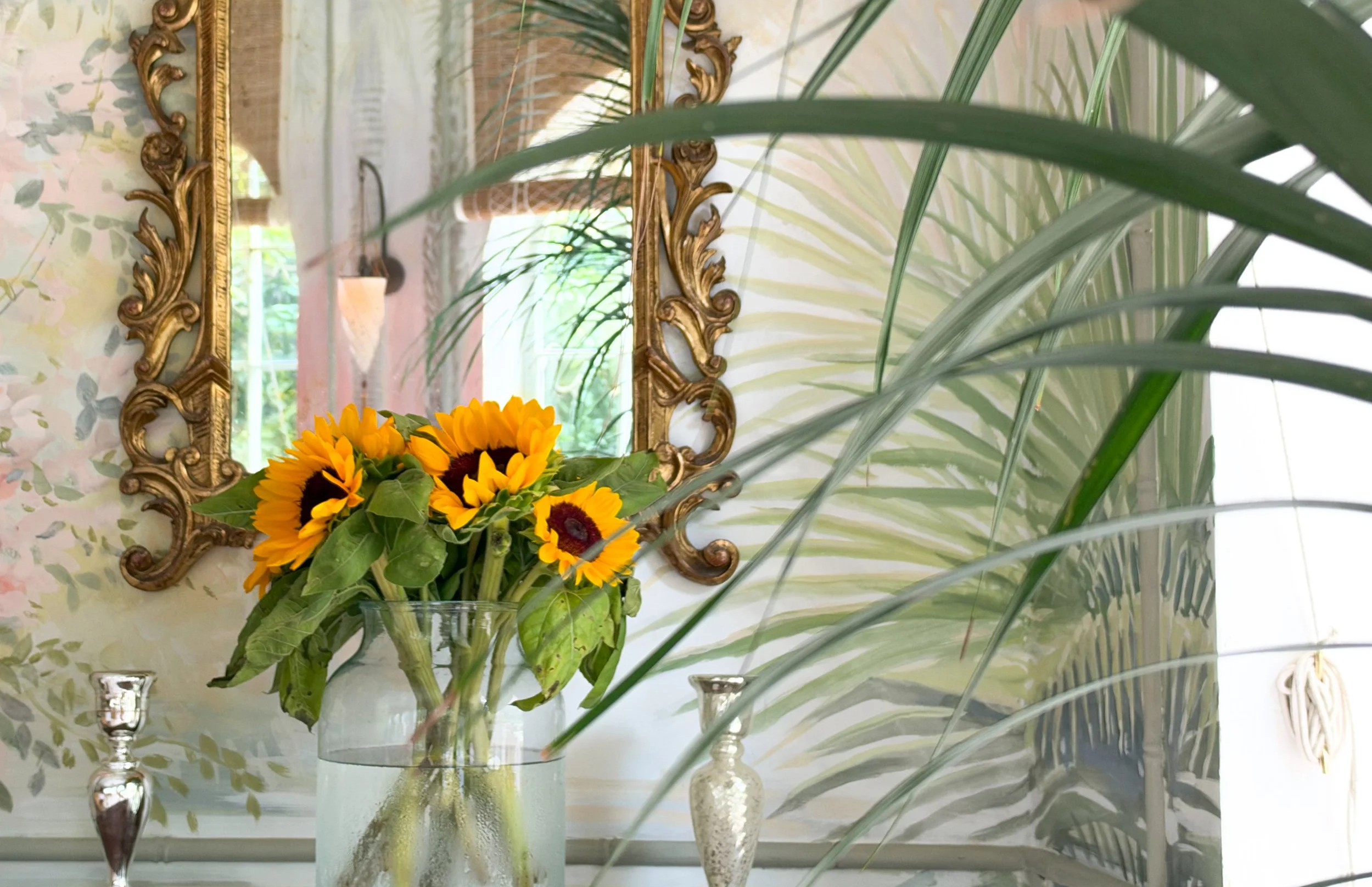 A floral arrangement of sunflowers in a glass vase with green leaves, placed on a table with three decorative candle holders. A gold ornate mirror hangs on a wall covered in pastel floral wallpaper, reflected in the mirror are green plants and windows.