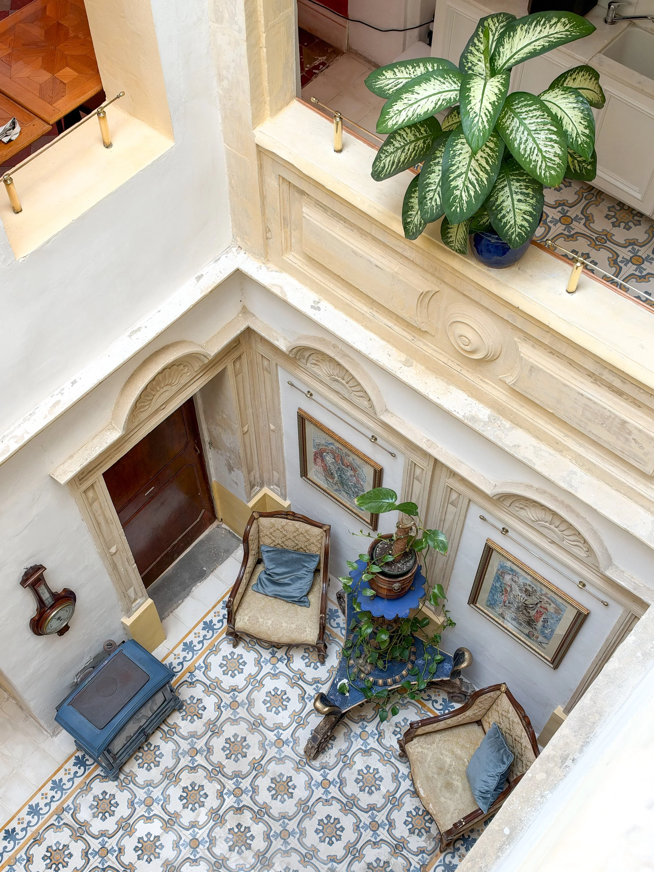 An interior view of a foyer with ornate tile flooring, vintage furniture, a large green potted plant, framed artwork, and vintage decorative items.