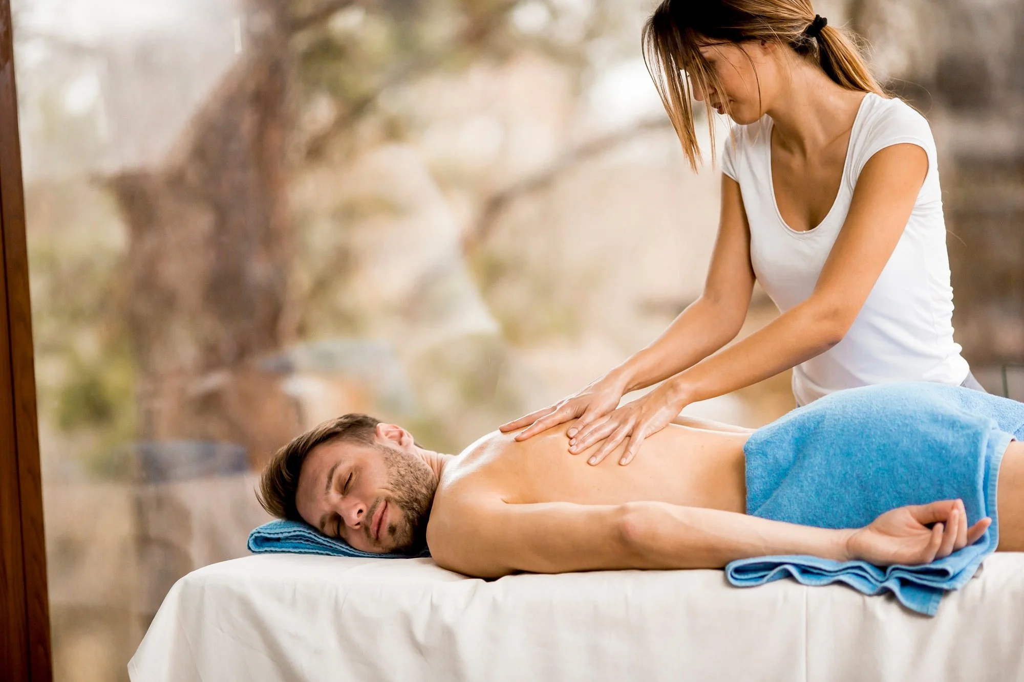 A woman giving a back massage to a man lying face down with eyes closed on a massage table. The woman is focused, wearing a white shirt, and the man is covered with a blue towel, in an outdoor natural setting with blurred trees in the background.