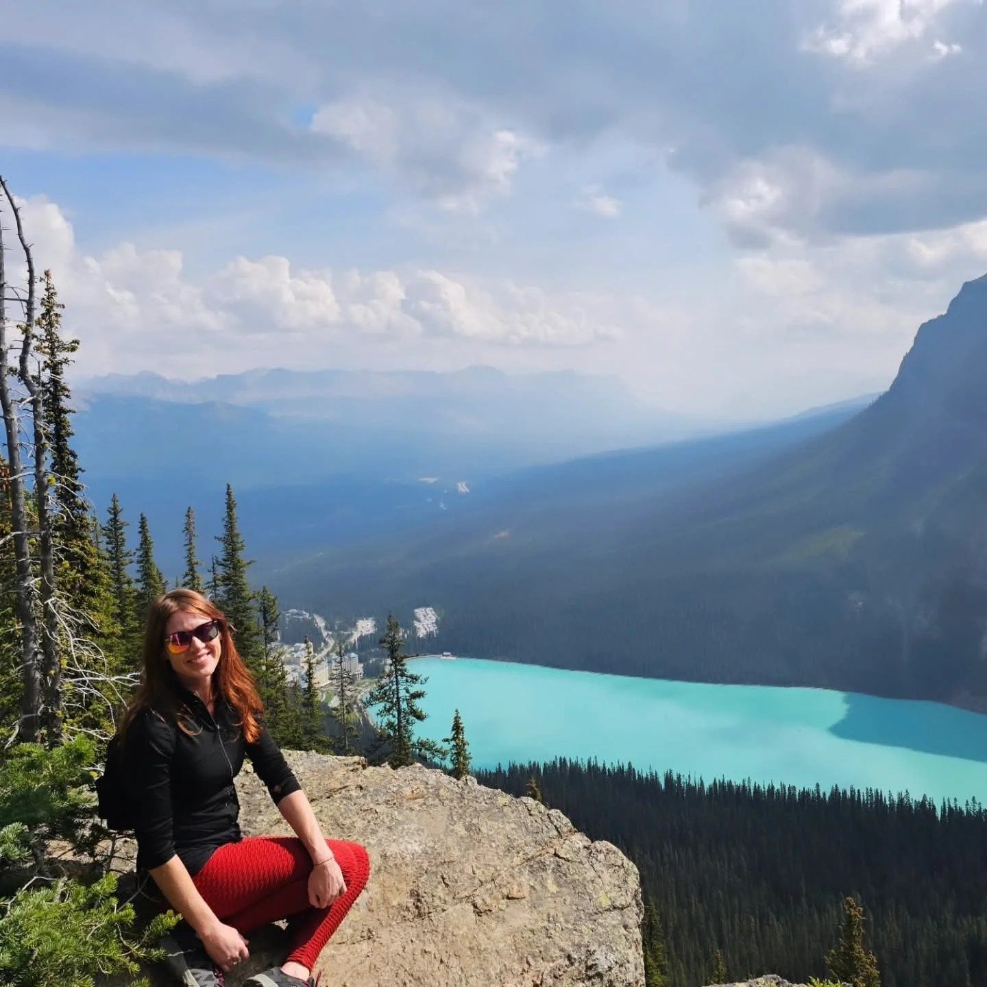 A woman with red hair, wearing sunglasses and a black jacket, sitting on a rock in front of a mountain landscape with a turquoise lake below.