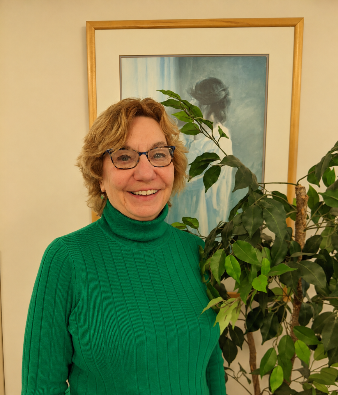 Smiling woman wearing green next to a plant in front of a painting