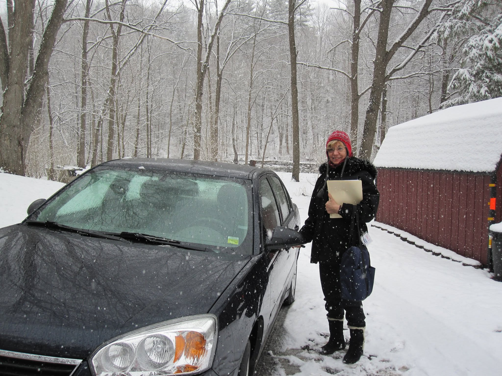 Woman in coat and hat in the snow next to a car and a barn