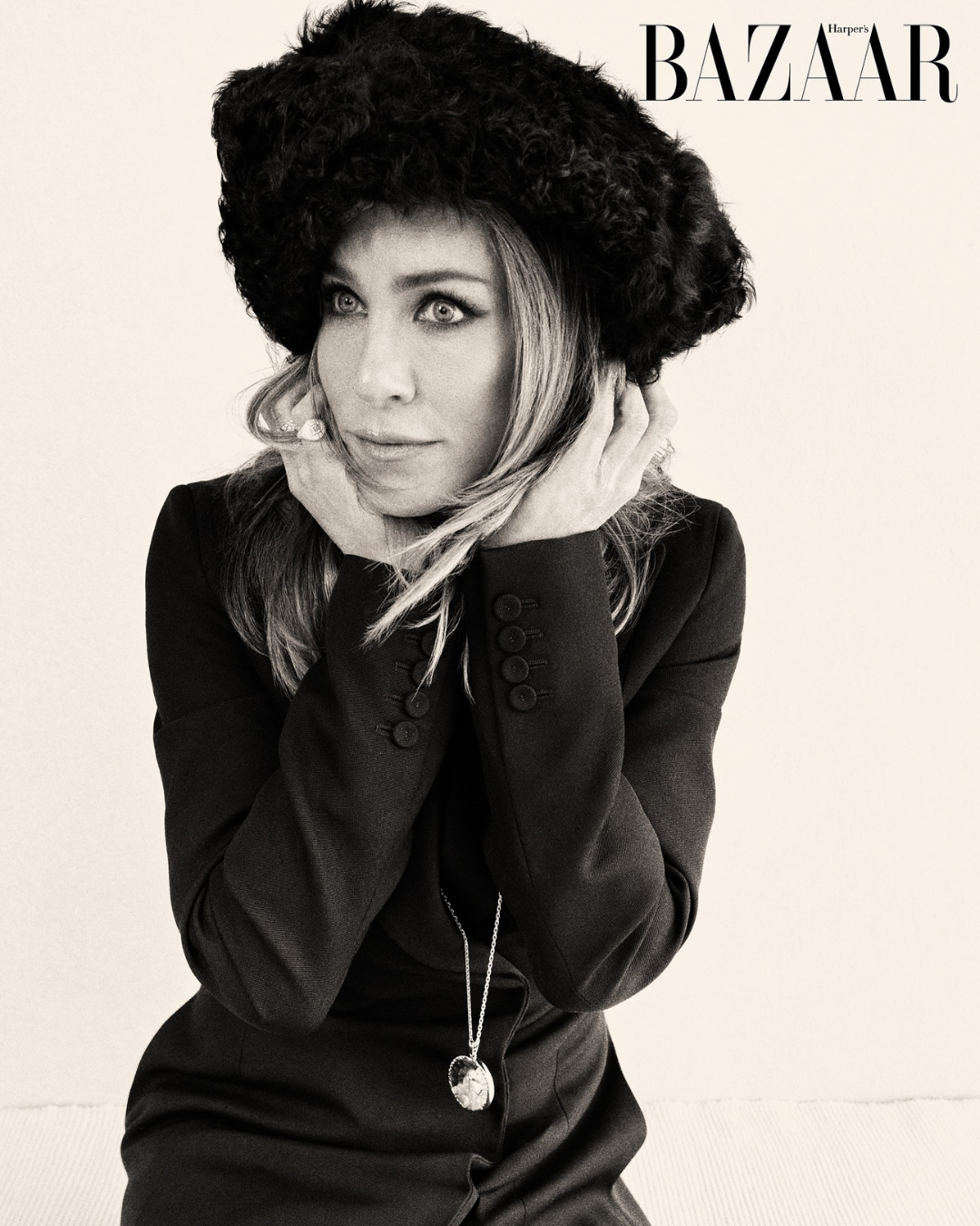 Black and white photo of a woman wearing a black blazer and a large black fuzzy hat, with her hands on her face, looking off to the side. The image has the Harper's Bazaar logo in the top right corner.