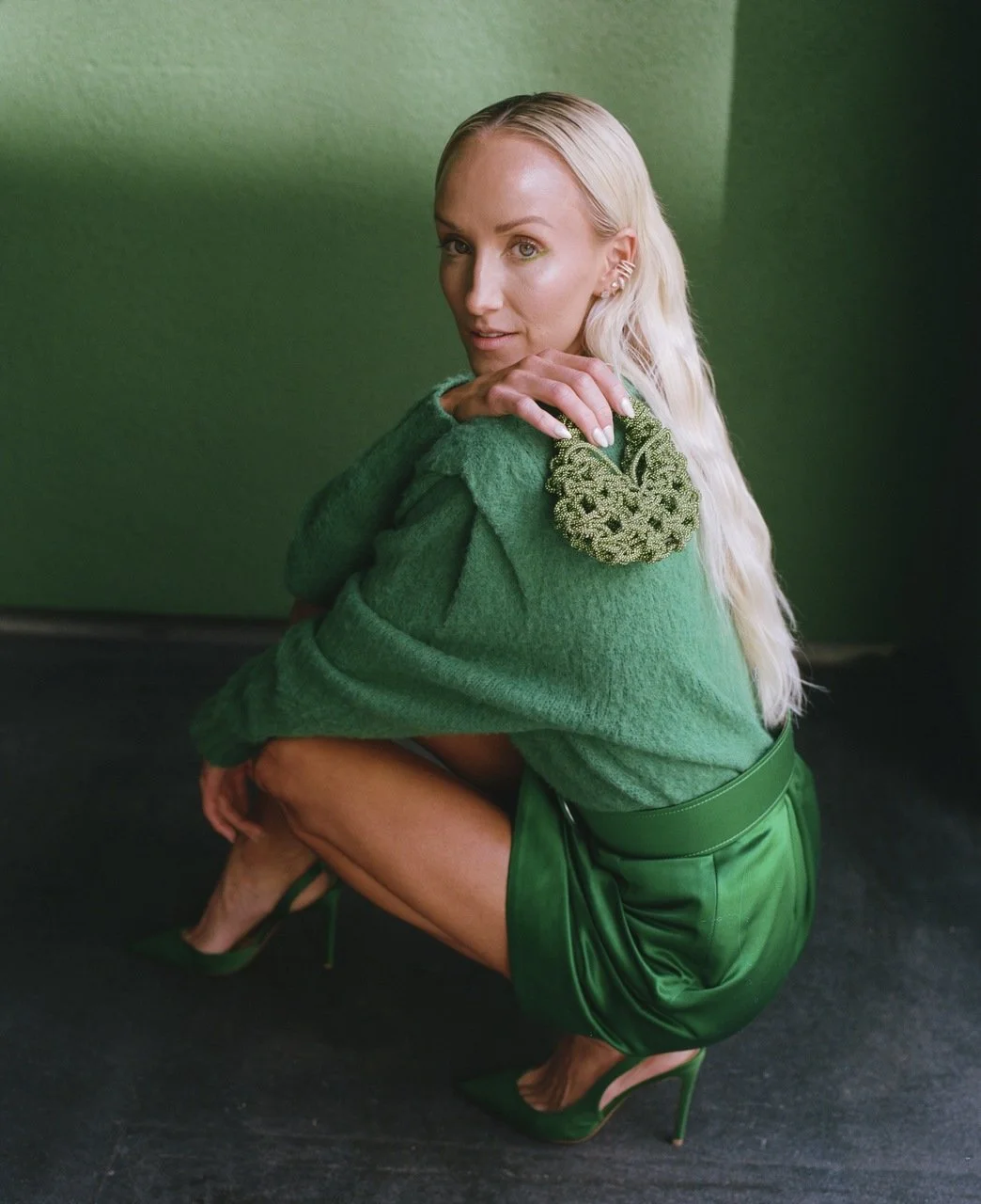 A woman with long blonde hair, wearing a green sweater with ornate embroidery on the shoulder, a green leather skirt, and green high heels, crouching against a green wall.