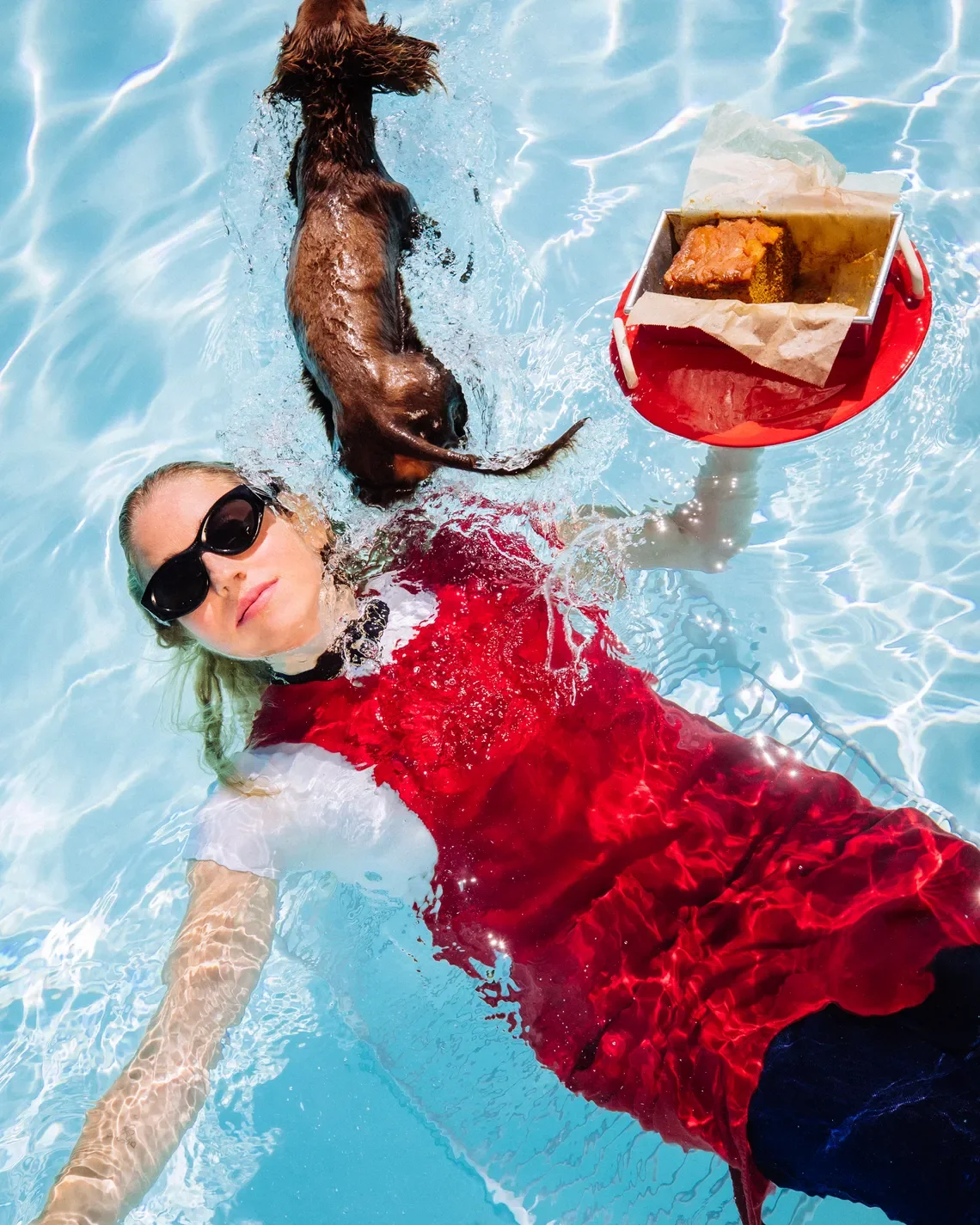A woman in sunglasses and a red dress swimming in a pool next to a brown dog, with a red tray holding a piece of cake floating nearby.