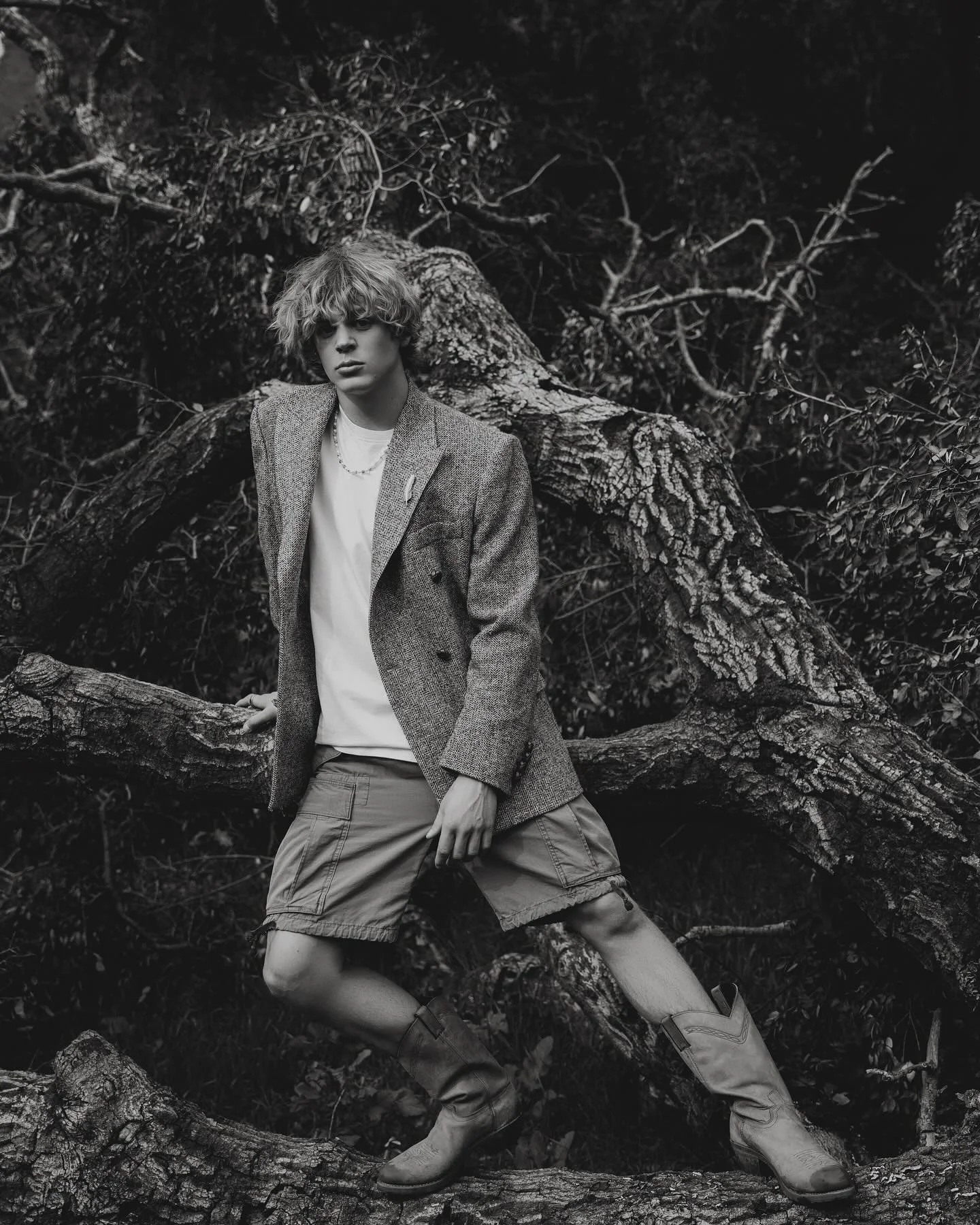Young man with curly hair wearing a blazer, t-shirt, cargo shorts, and boots standing among tree branches and fallen logs in a forest, in black and white.