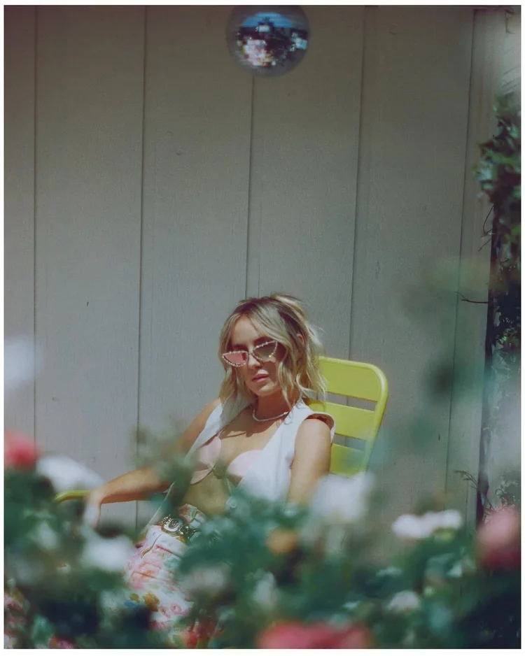 A woman with blonde wavy hair wearing pink sunglasses and a white sleeveless top, sitting on a yellow chair outdoors, surrounded by colorful flowers, with a beige wooden wall and small plant on the right side in the background, and a small disco ball