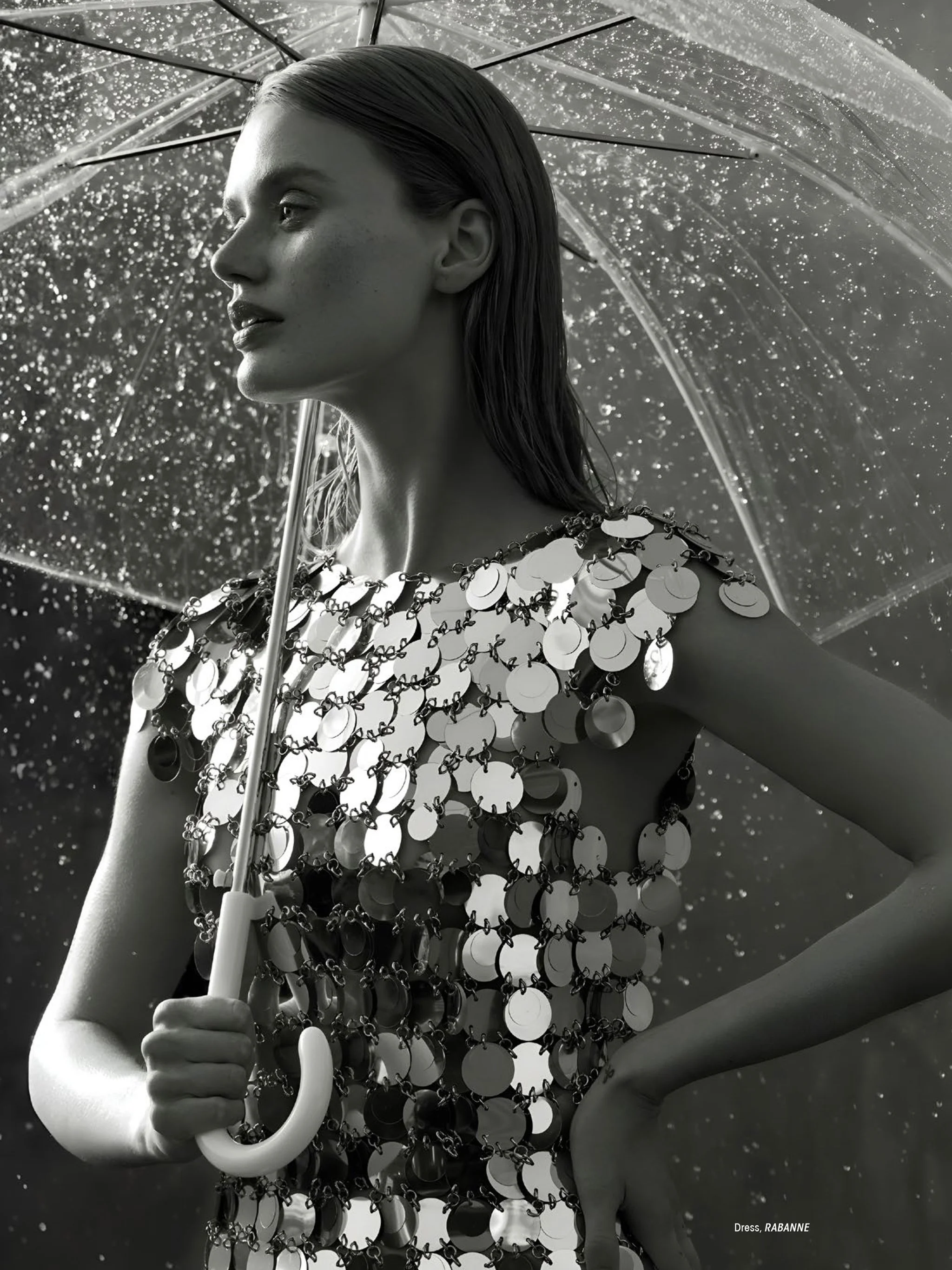 A black-and-white fashion photograph of a woman holding an umbrella in the rain. She is wearing a metallic, circular sequined dress by RABANNE and has her wet hair slicked back. The background is filled with raindrops.