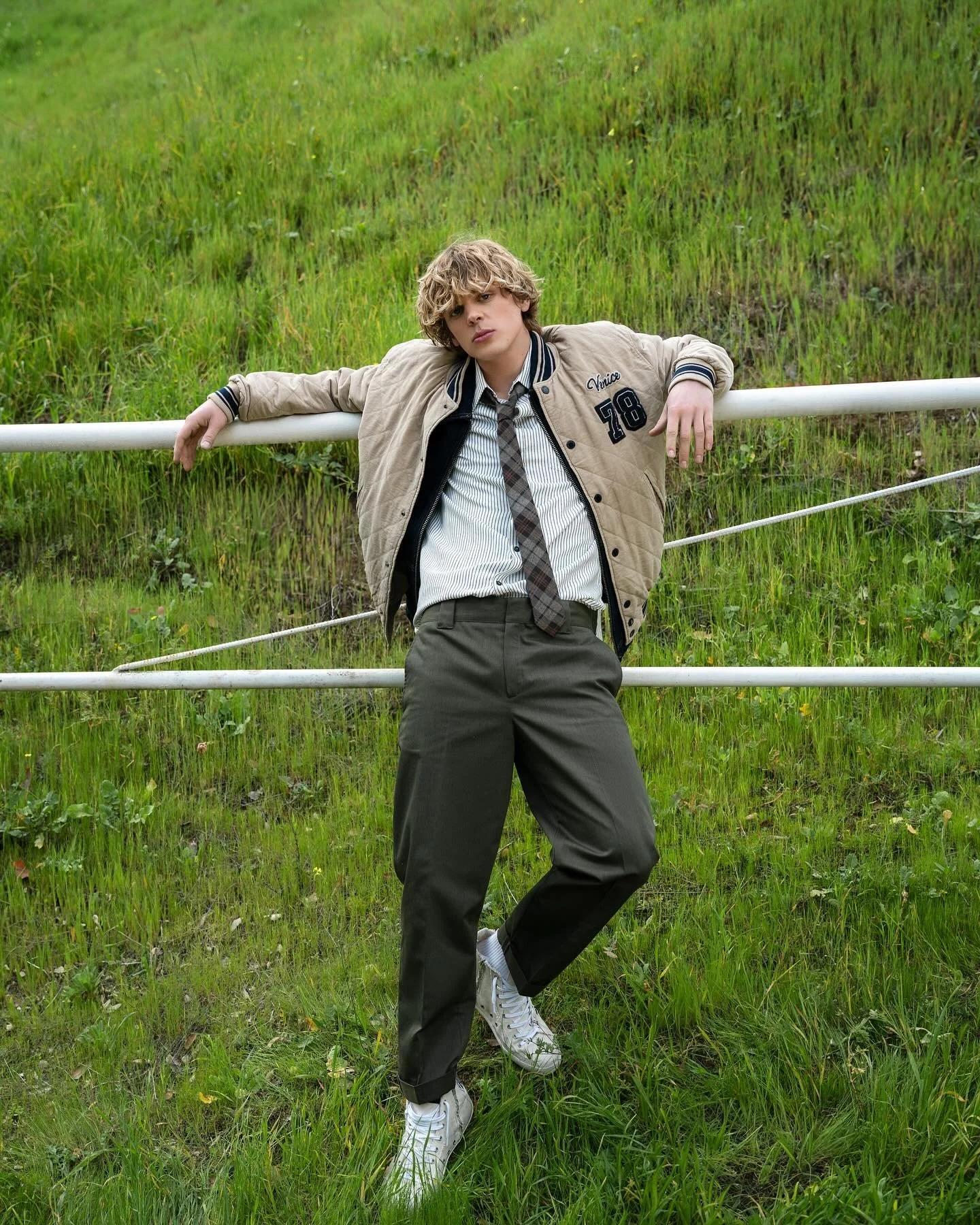 A young man with curly blond hair standing against a metal fence in a grassy field, wearing a beige jacket with patches, a striped shirt, a plaid tie, dark pants, and white sneakers, with his arms resting on the fence.