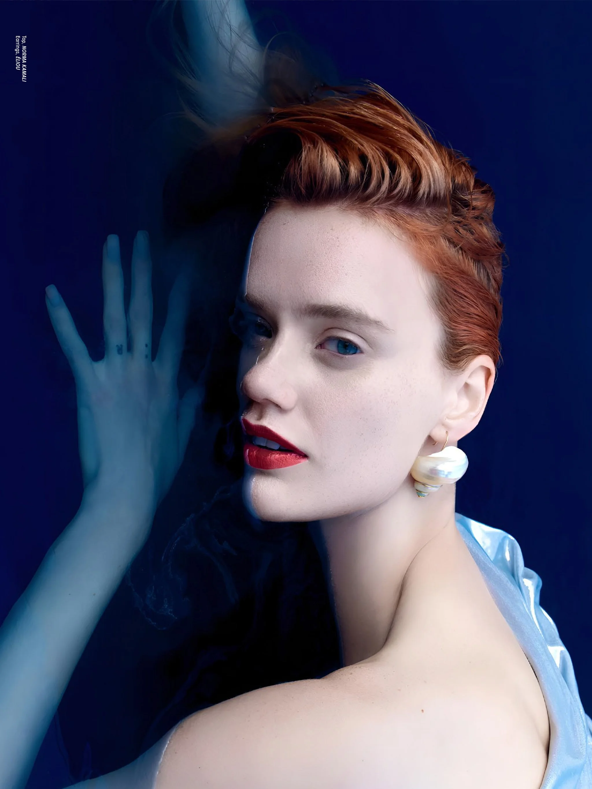 A woman with red hair styled in a vintage waves, wearing red lipstick, large pearl earring, and a light blue satin top, posed against a dark blue background with her hand near her face.