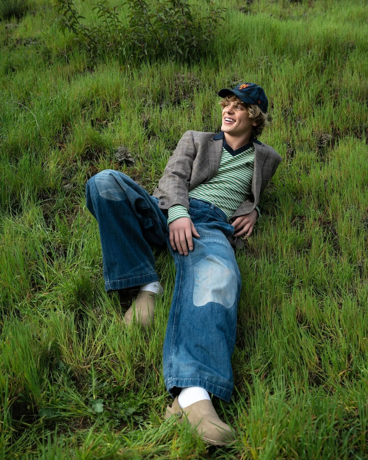A young man with light brown hair wearing a navy baseball cap, beige blazer, striped green and white shirt, and blue jeans, lying on the grass in a natural outdoor setting with green grass and some small bushes, smiling and looking happy.