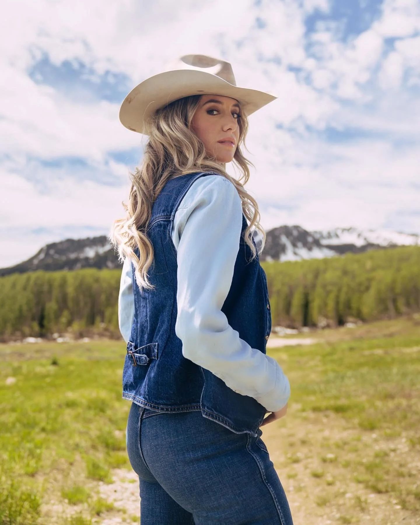 A woman with long blonde wavy hair wearing a cowboy hat, denim jacket over a light shirt, and jeans, standing outdoors in a field with mountains and trees in the background on a cloudy day.