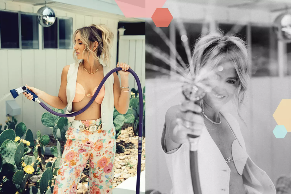 Side view of a young woman with blonde hair holding a garden hose and a spray gun, wearing a floral pants, peach-colored bikini top and sleeveless vest, standing outdoors with greenery and a white wooden fence.