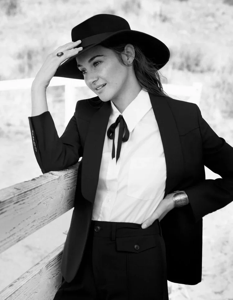 Black and white photo of a woman in a blazer, white shirt with a black ribbon tie, and a wide-brimmed hat, leaning on a wooden fence outdoors.