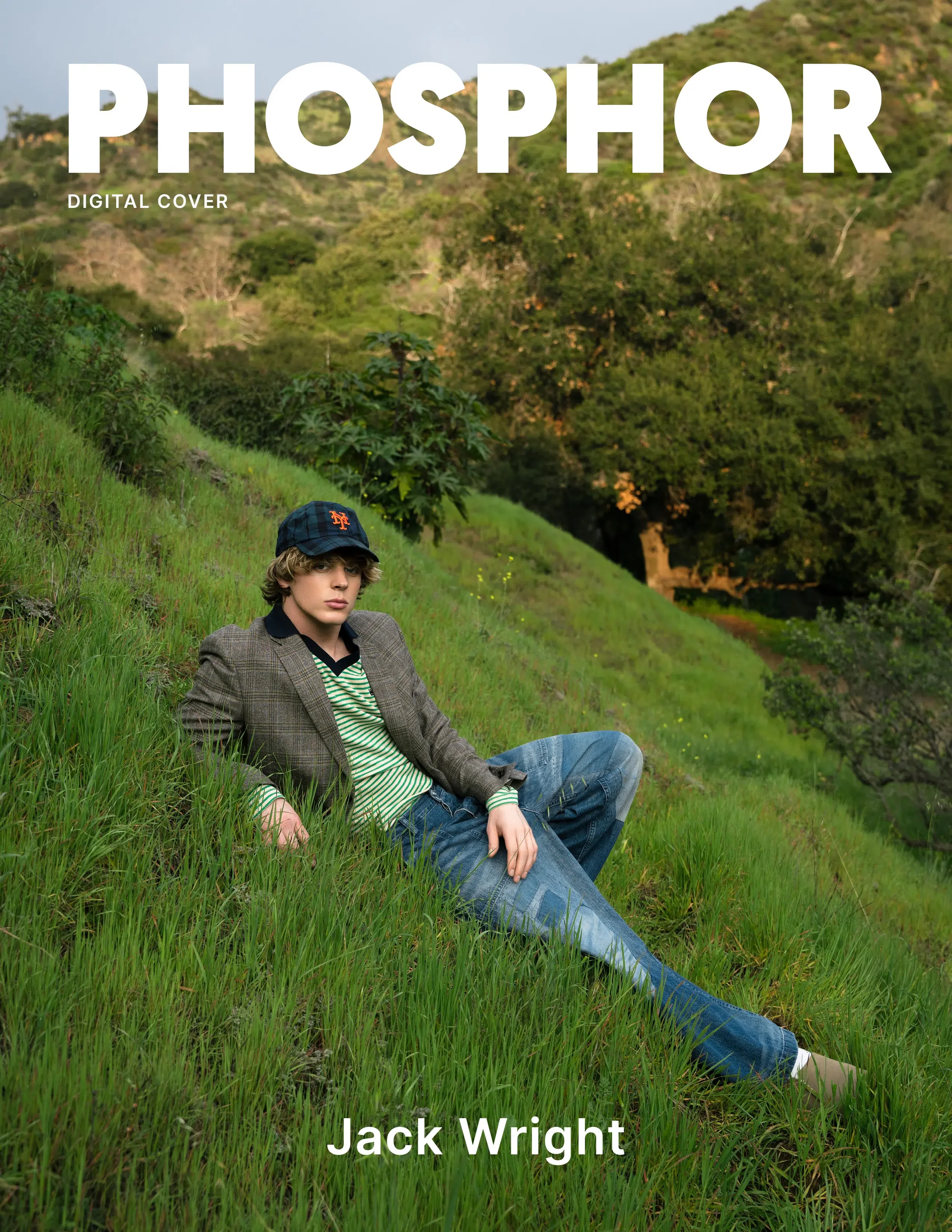 Young man with blond hair wearing a baseball cap and a plaid blazer over a striped shirt, sitting on grassy hillside with trees and hills in background, facing slightly left.