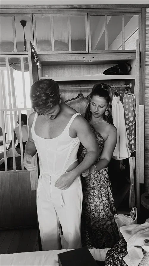 A woman helping a woman try on a dress in a bedroom, with clothes hanging in the closet behind them.