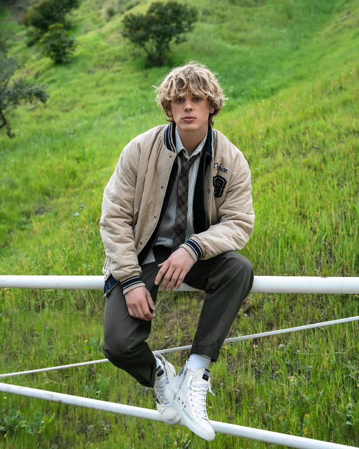 Young man with curly blonde hair sitting on a white fence in a grassy, hilly outdoor area with small green trees.