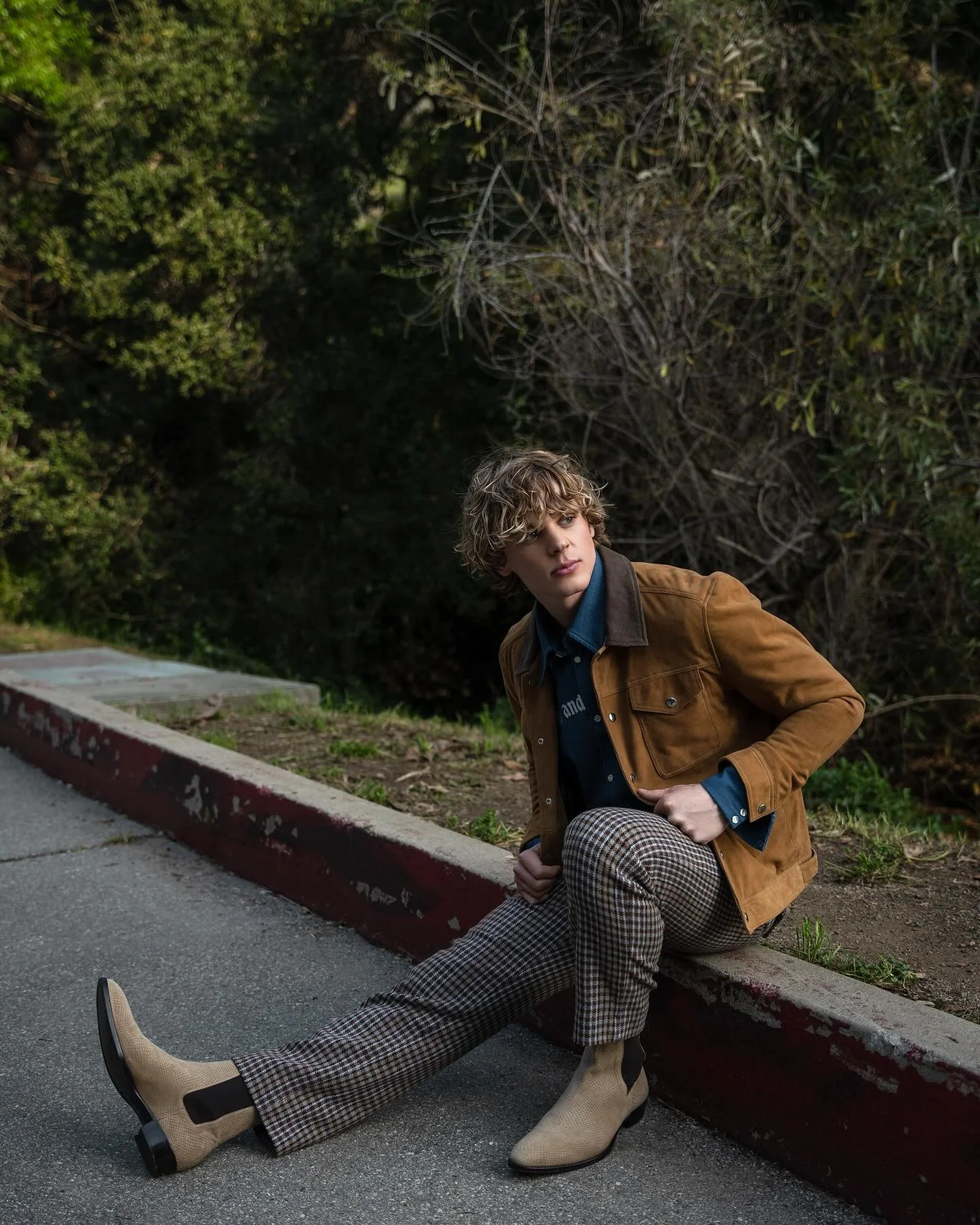 A person with curly hair wearing a brown jacket, plaid pants, and beige boots sitting on a curb next to a sidewalk with a background of trees and bushes.