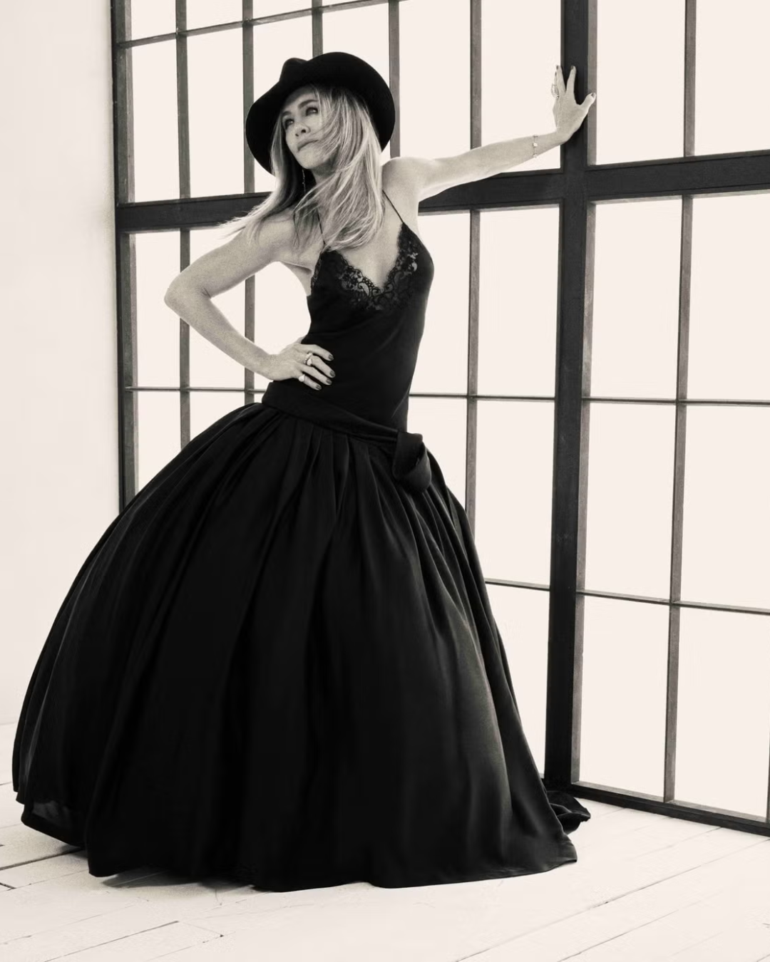 A woman in a black hat, lace top, and large black skirt posing with one hand on her hip and the other on a wall, standing in front of a large window with grid panes.