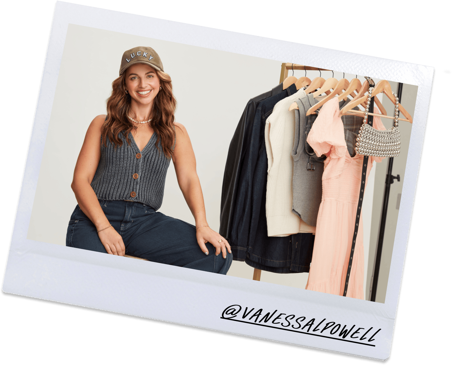 A woman smiling, sitting next to a clothing rack with various garments including jackets, sweaters, a dress, and a beaded handbag.