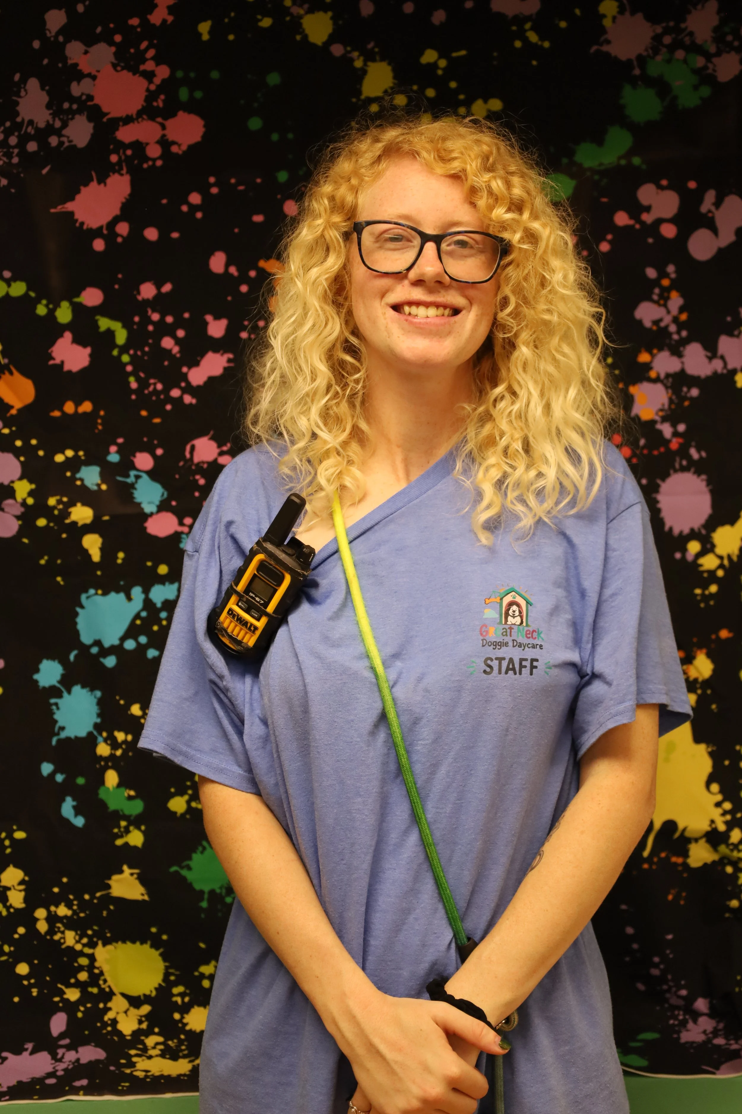 A smiling woman with curly blonde hair, wearing glasses and a blue T-shirt with a dog daycare logo, standing in front of a black background with colorful paint splatters. She has a walkie-talkie clipped to her shirt and a green leash hanging from her neck.
