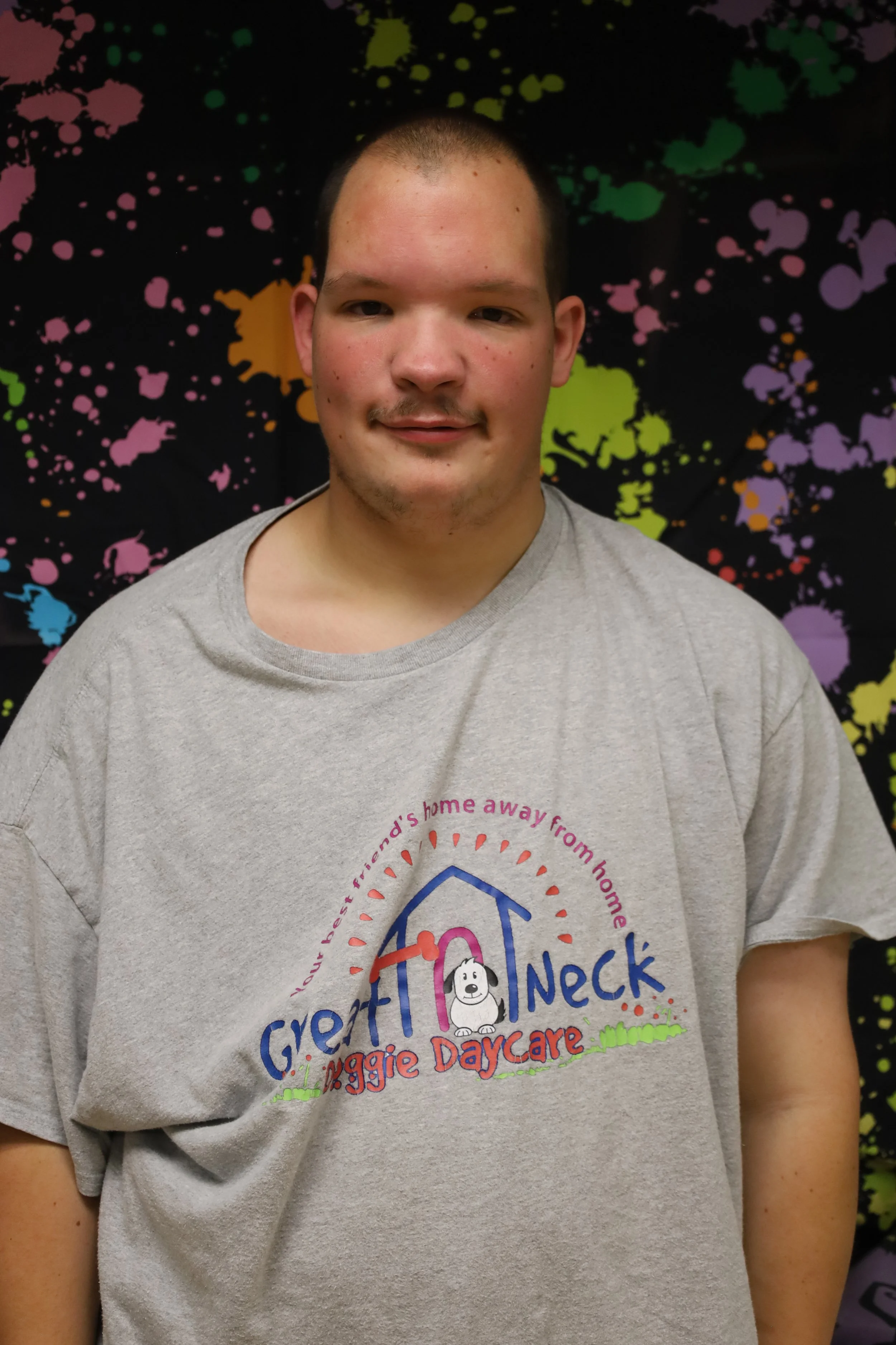 Young man wearing a gray t-shirt with a colorful logo and text related to Dog Rescue Support at a daycare, standing in front of a black background with multicolored paint splatter design.