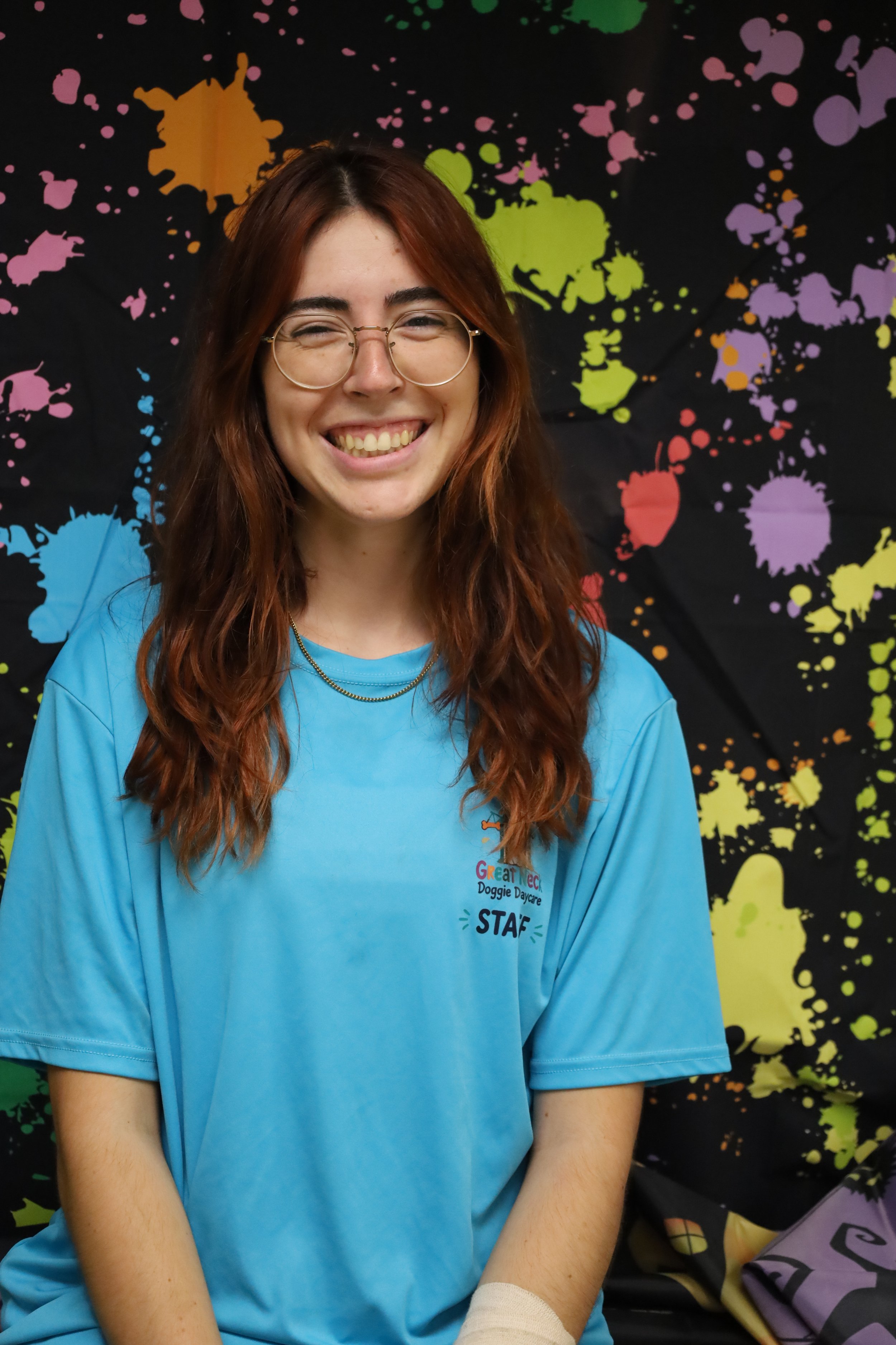 A smiling young woman with long wavy reddish-brown hair, wearing glasses and a blue T-shirt with a logo that reads 'Great Pet Doggie Daycare STAFF', sitting in front of a colorful splatter paint background.