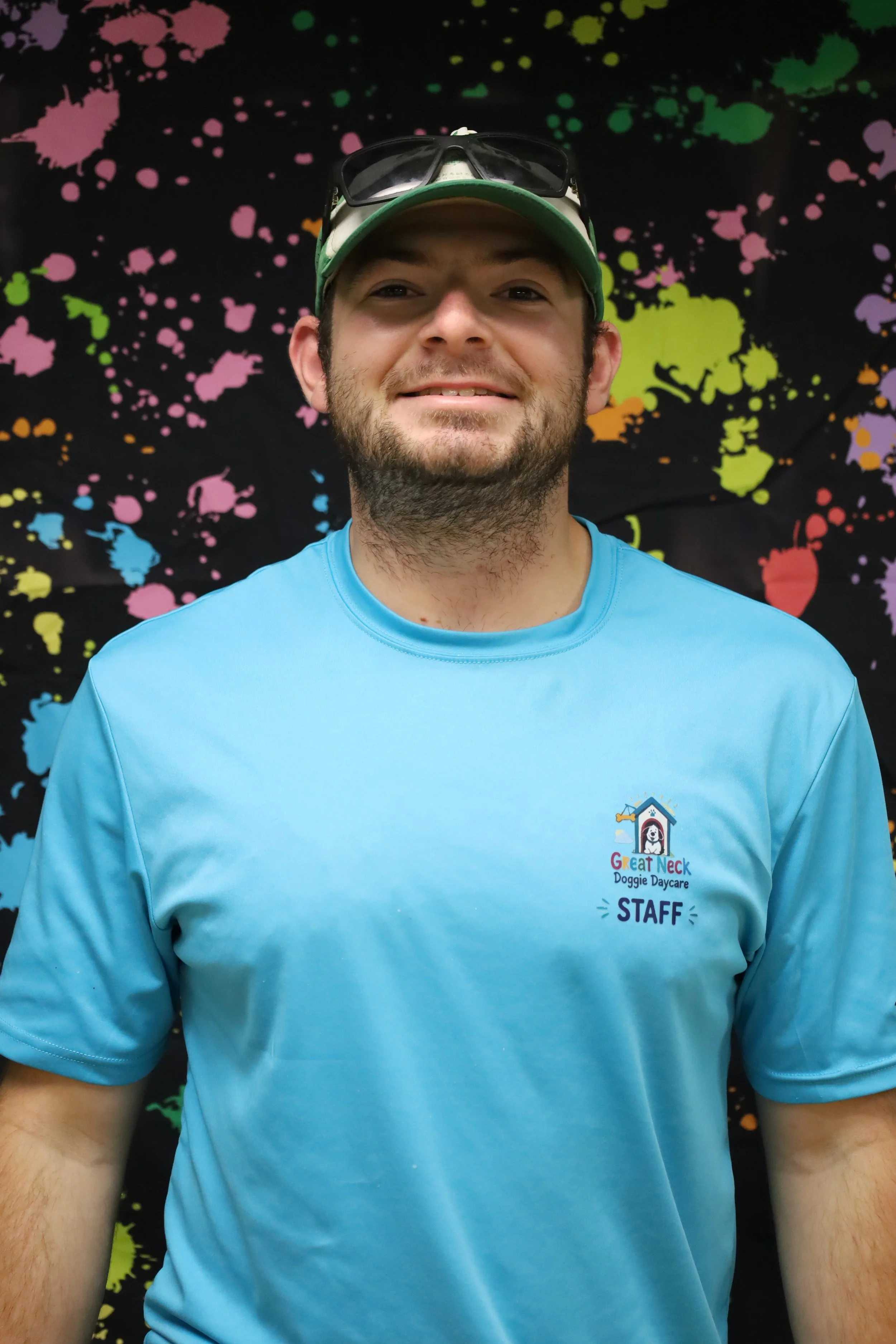 A smiling man wearing a green and white cap with sunglasses on top, blue t-shirt with a logo that says "Great Neck Doggie Daycare STAFF," against a colorful, splattered paint backdrop.