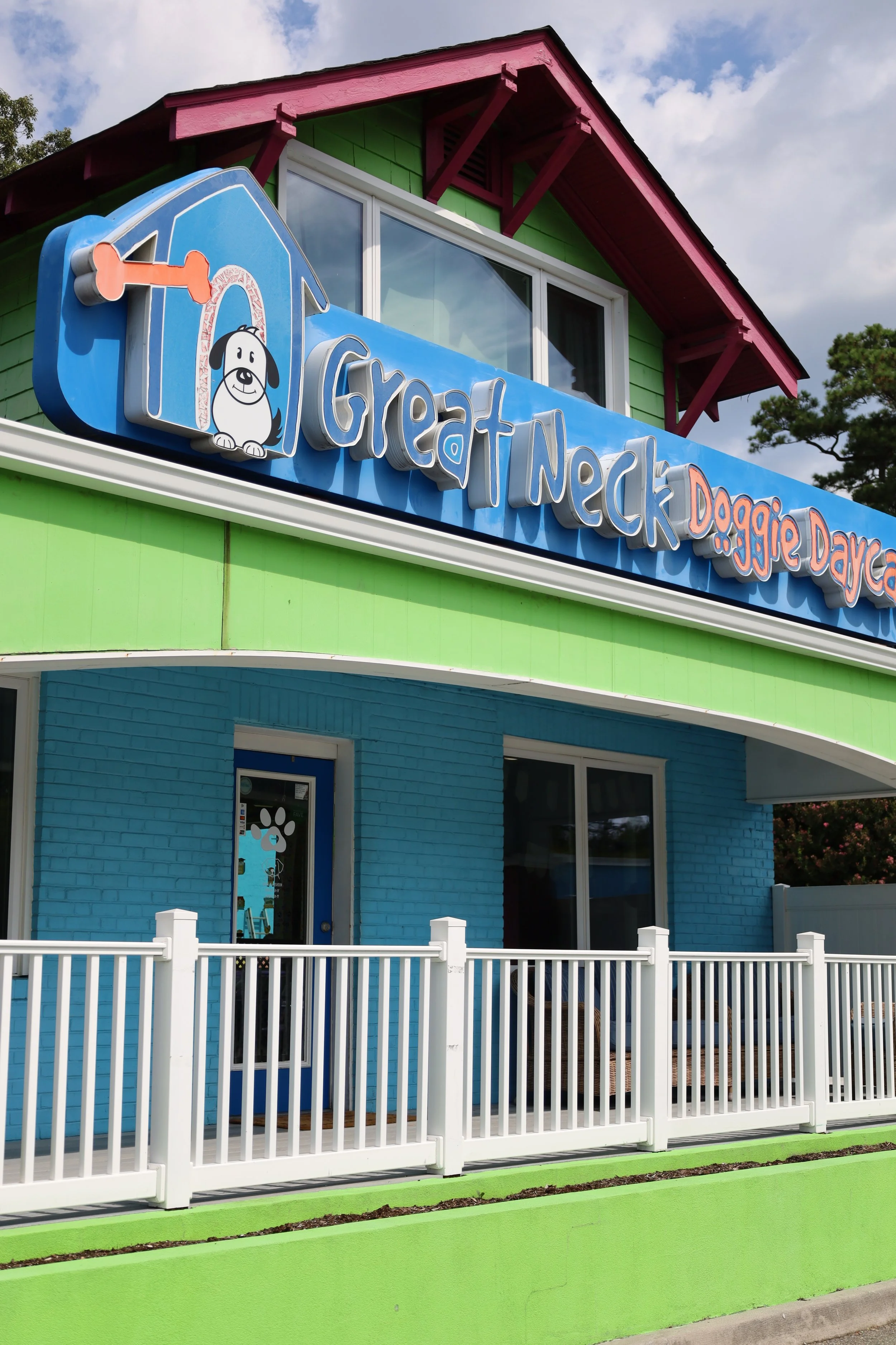 Colorful building with a sign reading 'Great Neck Doggie Daycare' featuring a cartoon dog and house. The building has a bright green lower section, blue middle section, and a pink upper roof. White fence in front.