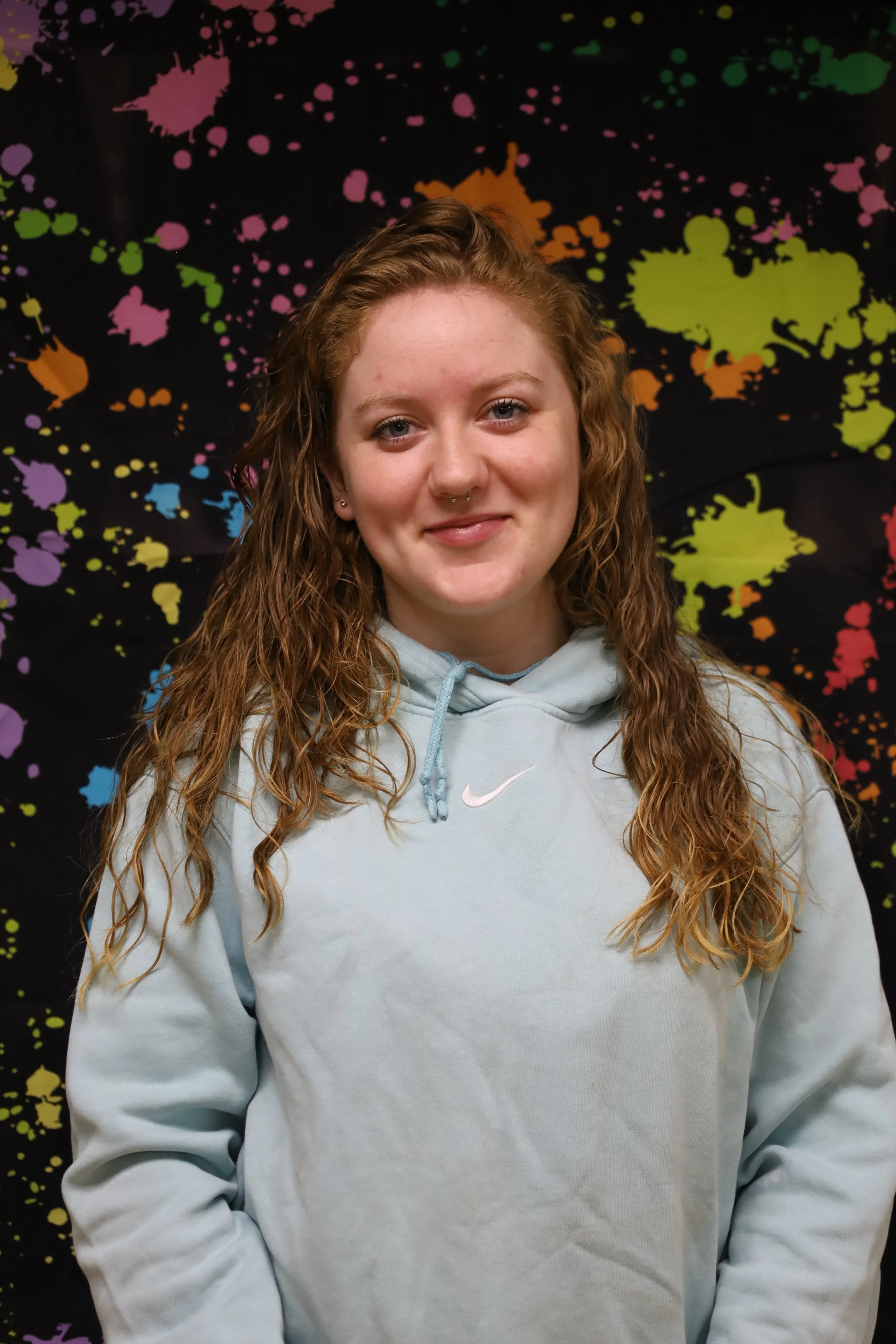 A young woman with long, curly red hair and light skin, wearing a light blue hoodie with a white Nike logo, stands in front of a colorful splatter paint backdrop.