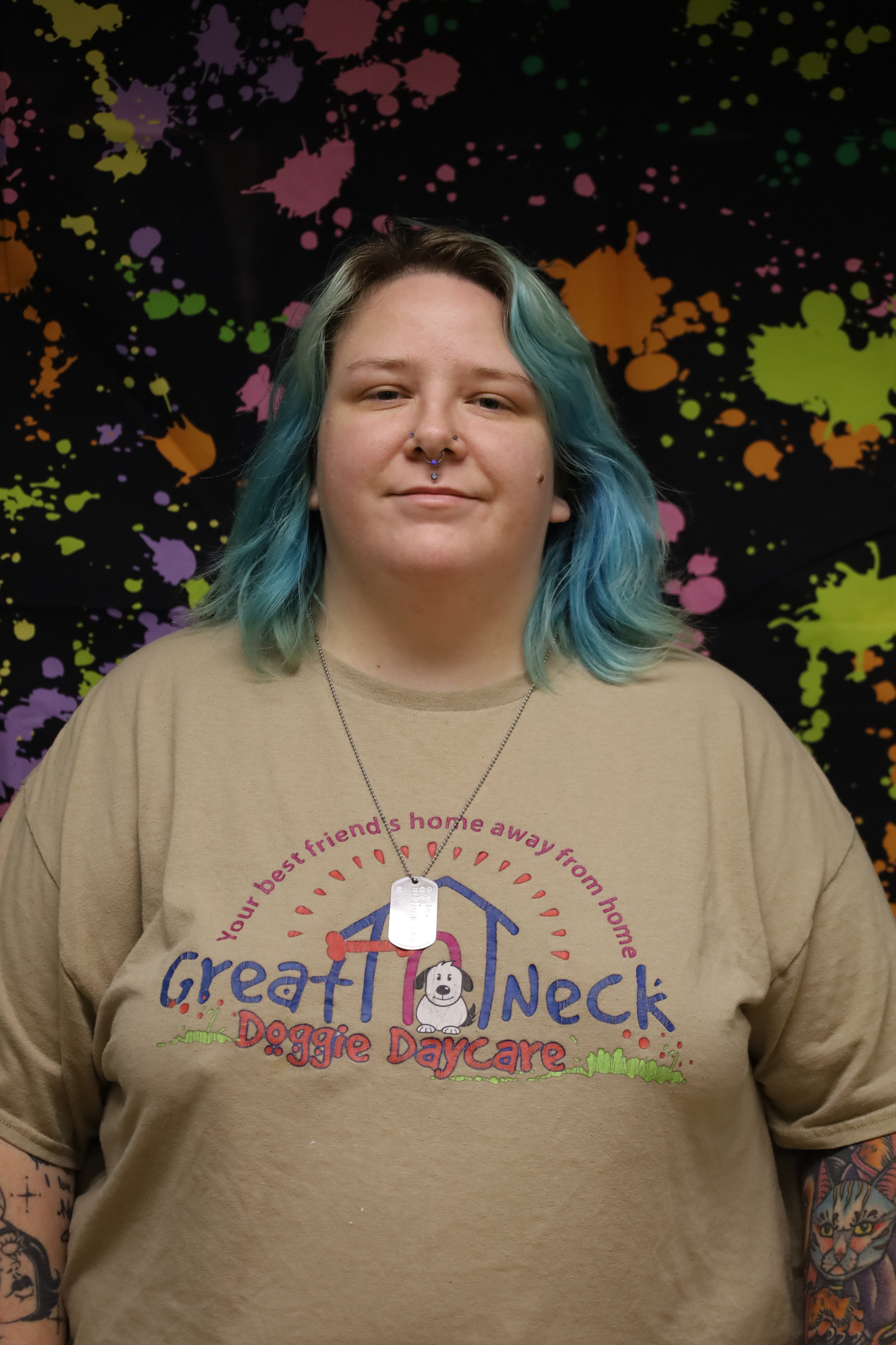 Person with blue-green hair wearing a beige sweatshirt featuring a colorful logo with text and graphics, standing in front of a black background with colorful paint splatter patterns.