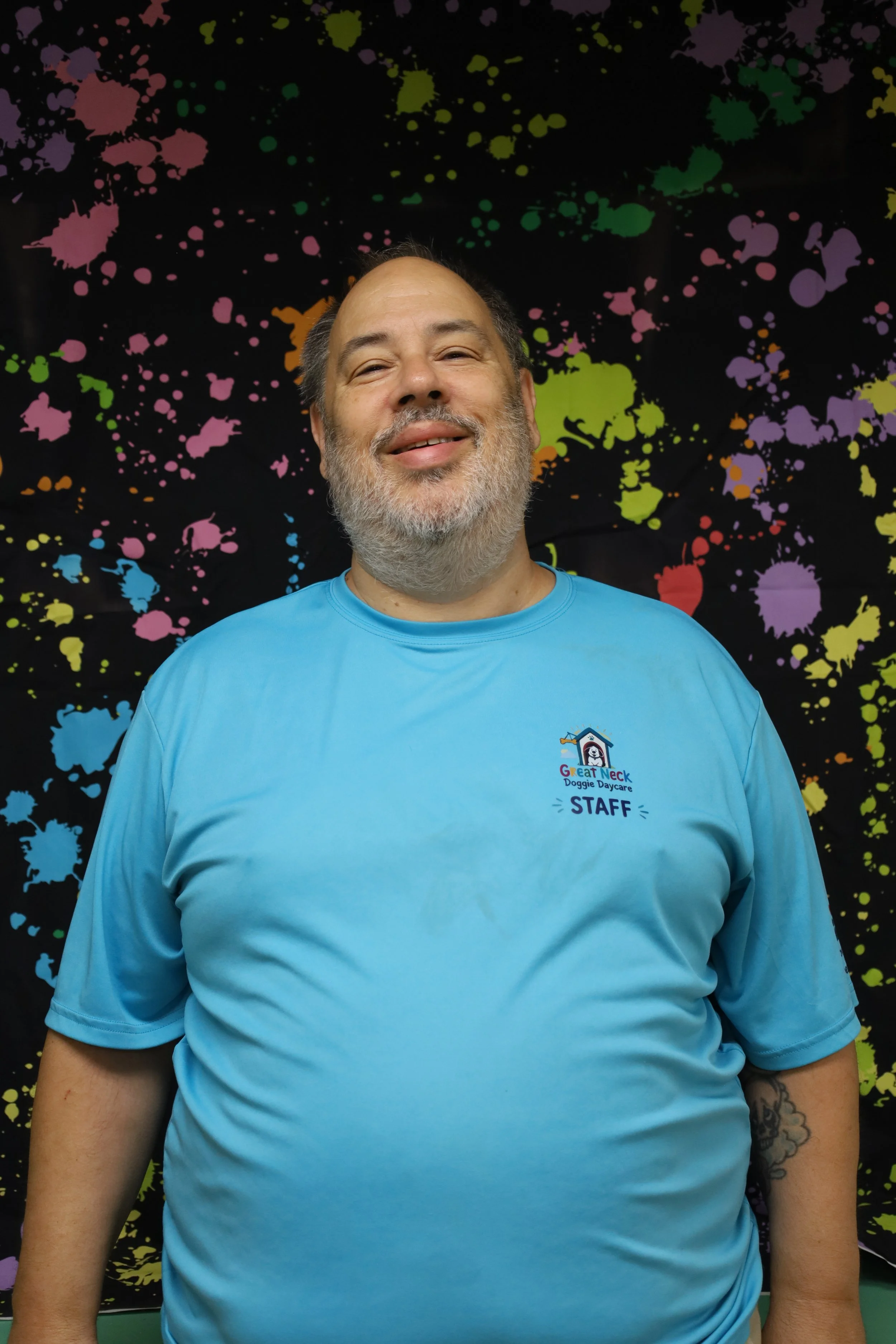 A middle-aged man with a beard and mustache, wearing a light blue T-shirt with a logo and the word "STAFF" on it, standing in front of a black background with colorful paint splatters.