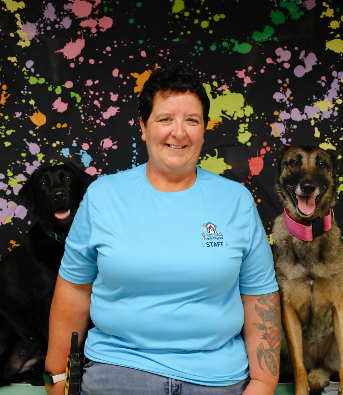 A smiling woman wearing a light blue shirt with a staff badge, sitting with two dogs in front of a colorful splatter paint backdrop.