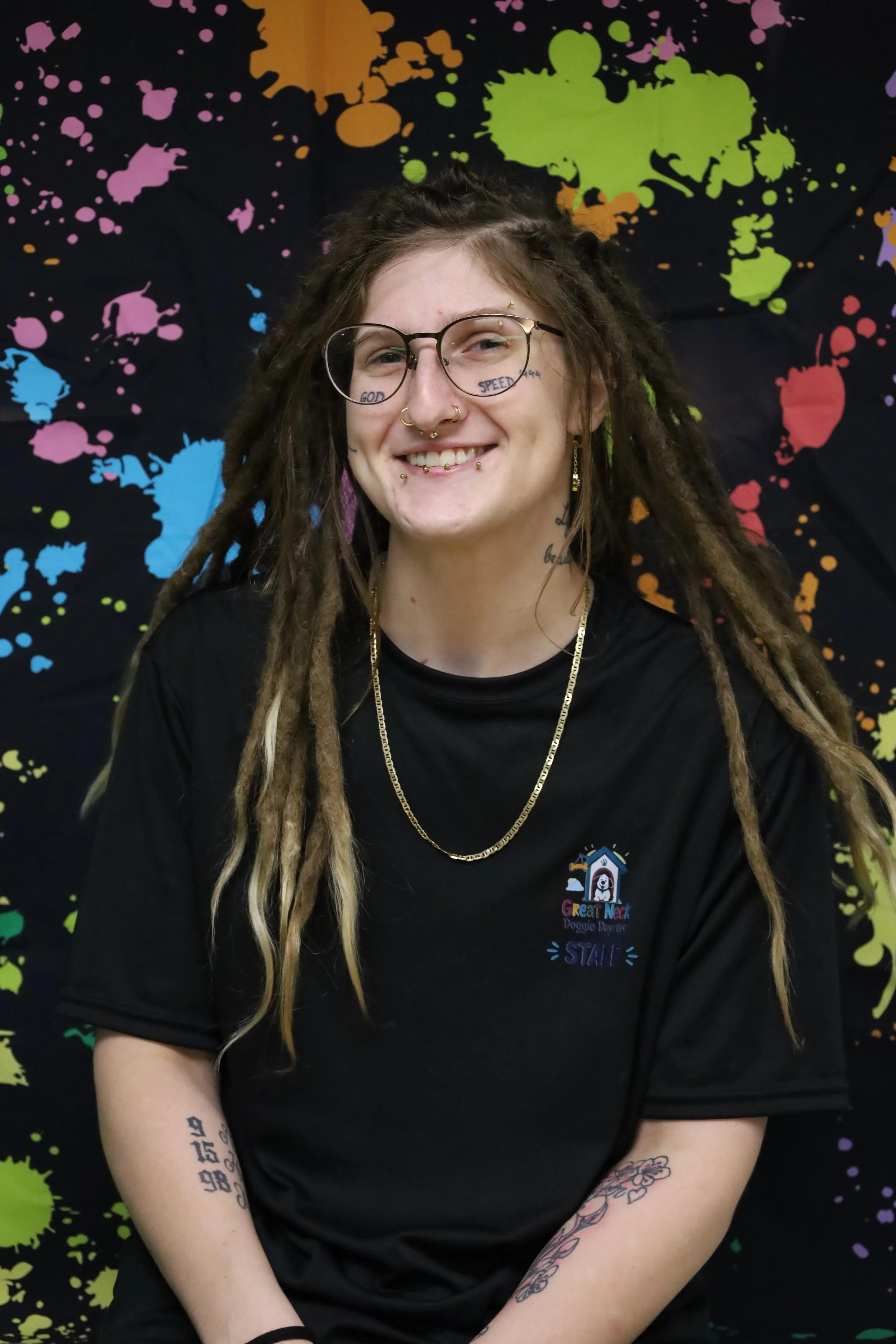 A smiling young woman with glasses, dreadlocks, and multiple tattoos on her arms, wearing a black T-shirt with a logo that says 'Great Neck Doggie Douter.' She is standing in front of a colorful, splatter-paint background.
