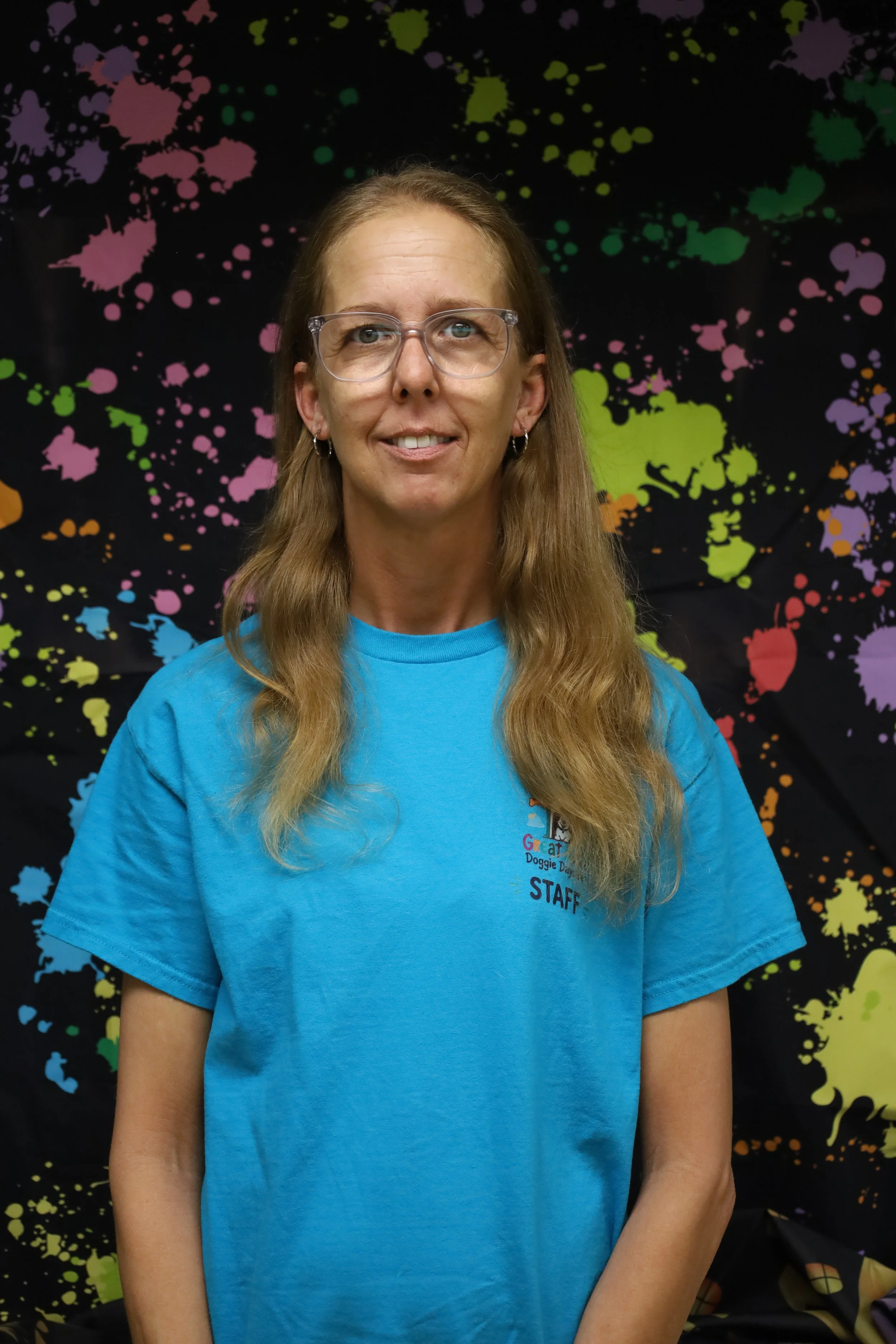 A woman with long blond hair, glasses, and earrings wearing a blue staff T-shirt, standing in front of a black background with colorful paint splatter patterns.